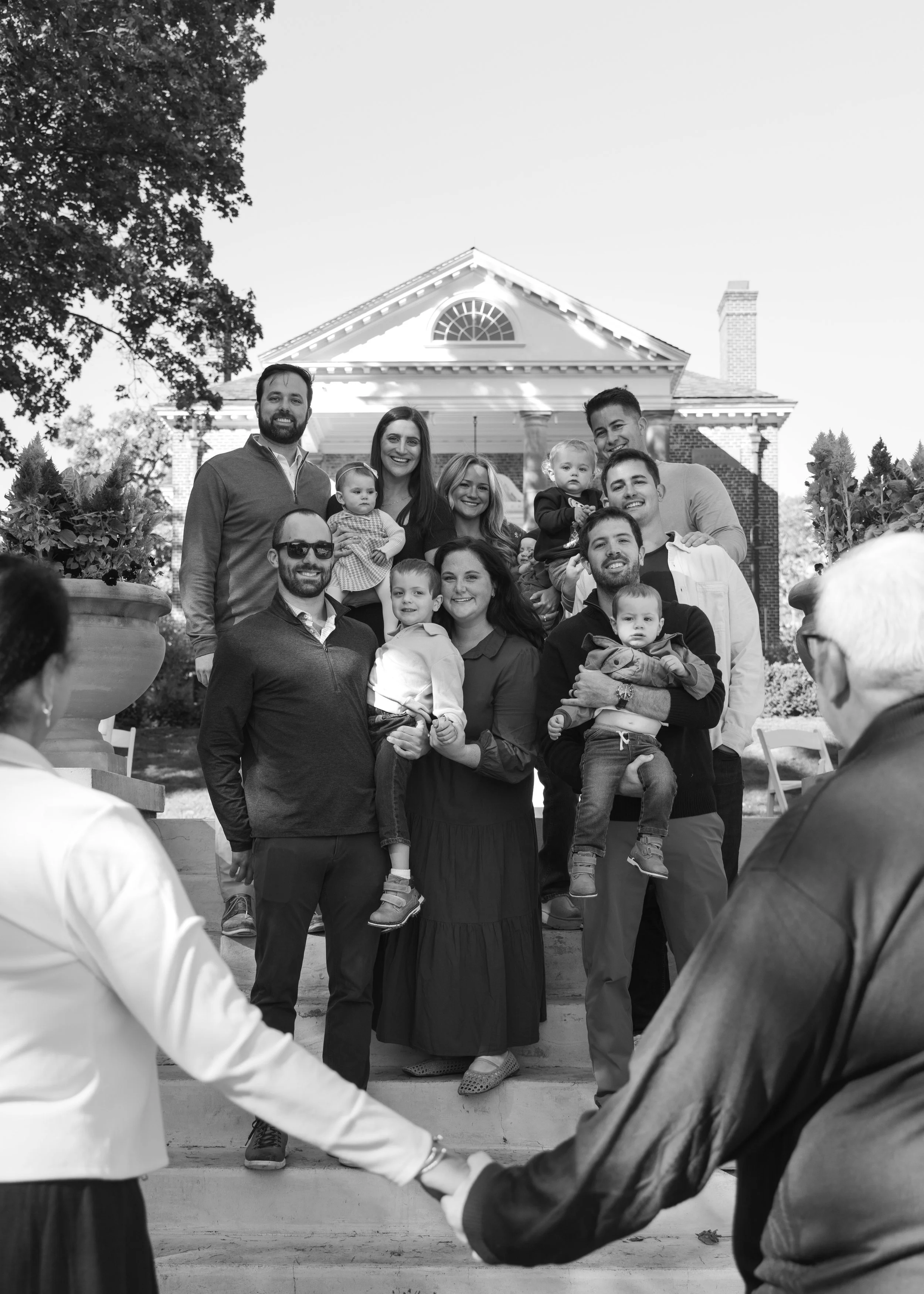Black and white photo of a large group of adults and children on the steps of a house, smiling and posing for a family photo, with two people holding hands at the front.
