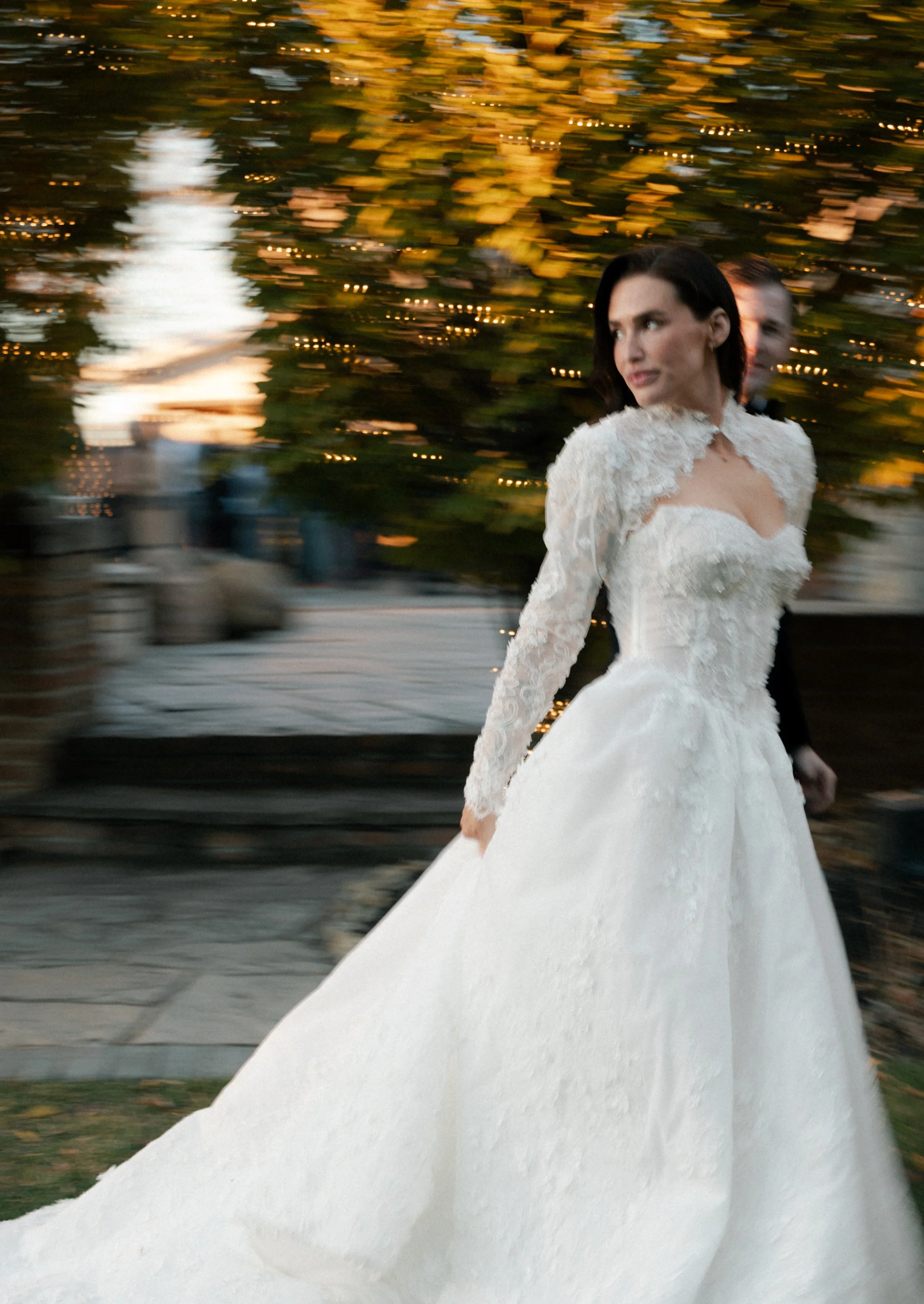 A woman in a white wedding dress walking outdoors, with blurred background of trees and string lights.