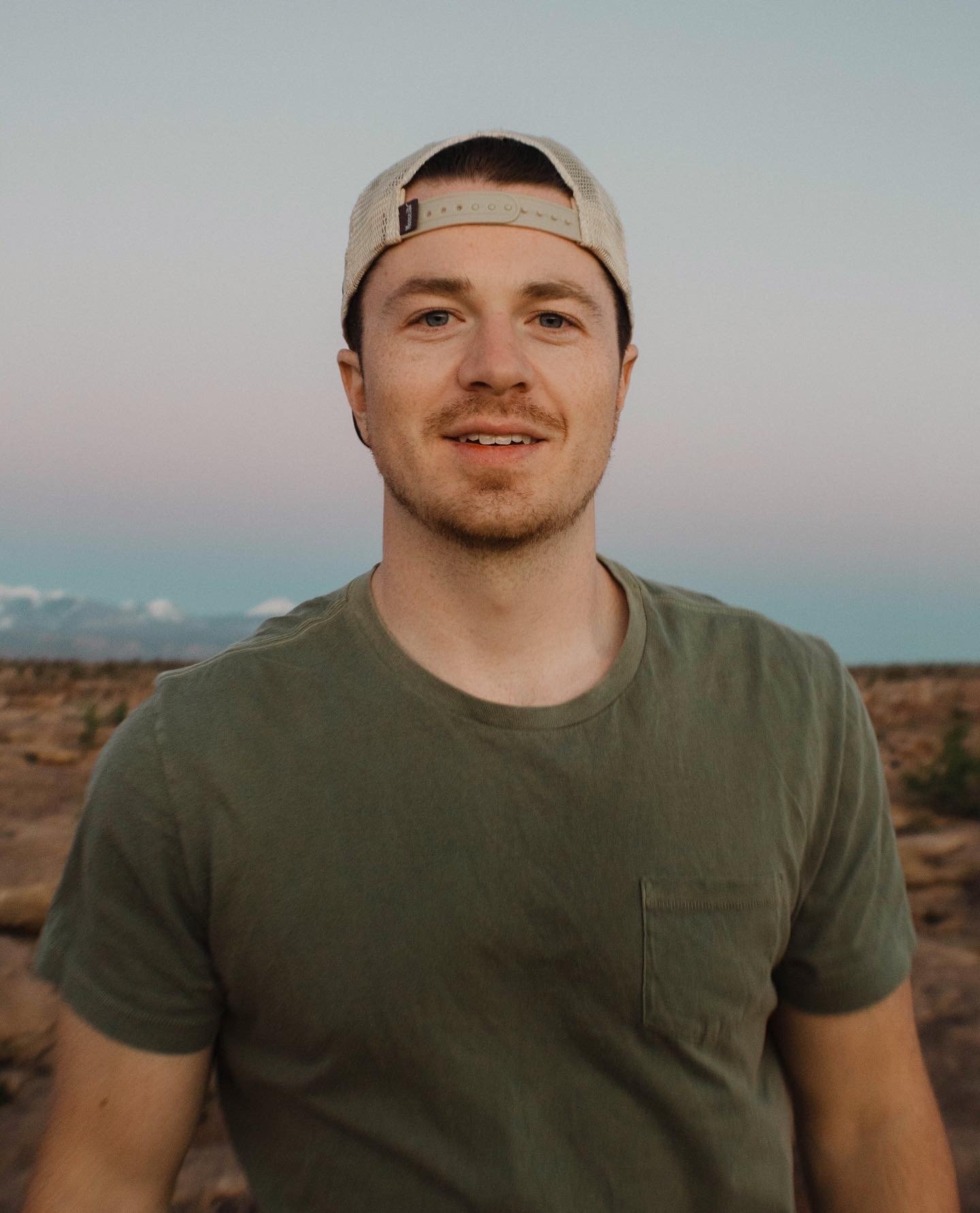 A young man smiling outdoors in a desert landscape at sunset, wearing a khaki t-shirt and a beige backward baseball cap.