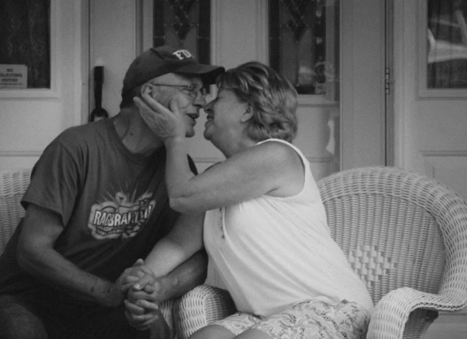 An elderly man and woman sit close together, touching foreheads and holding hands, sharing an intimate moment in a cozy indoor setting.
