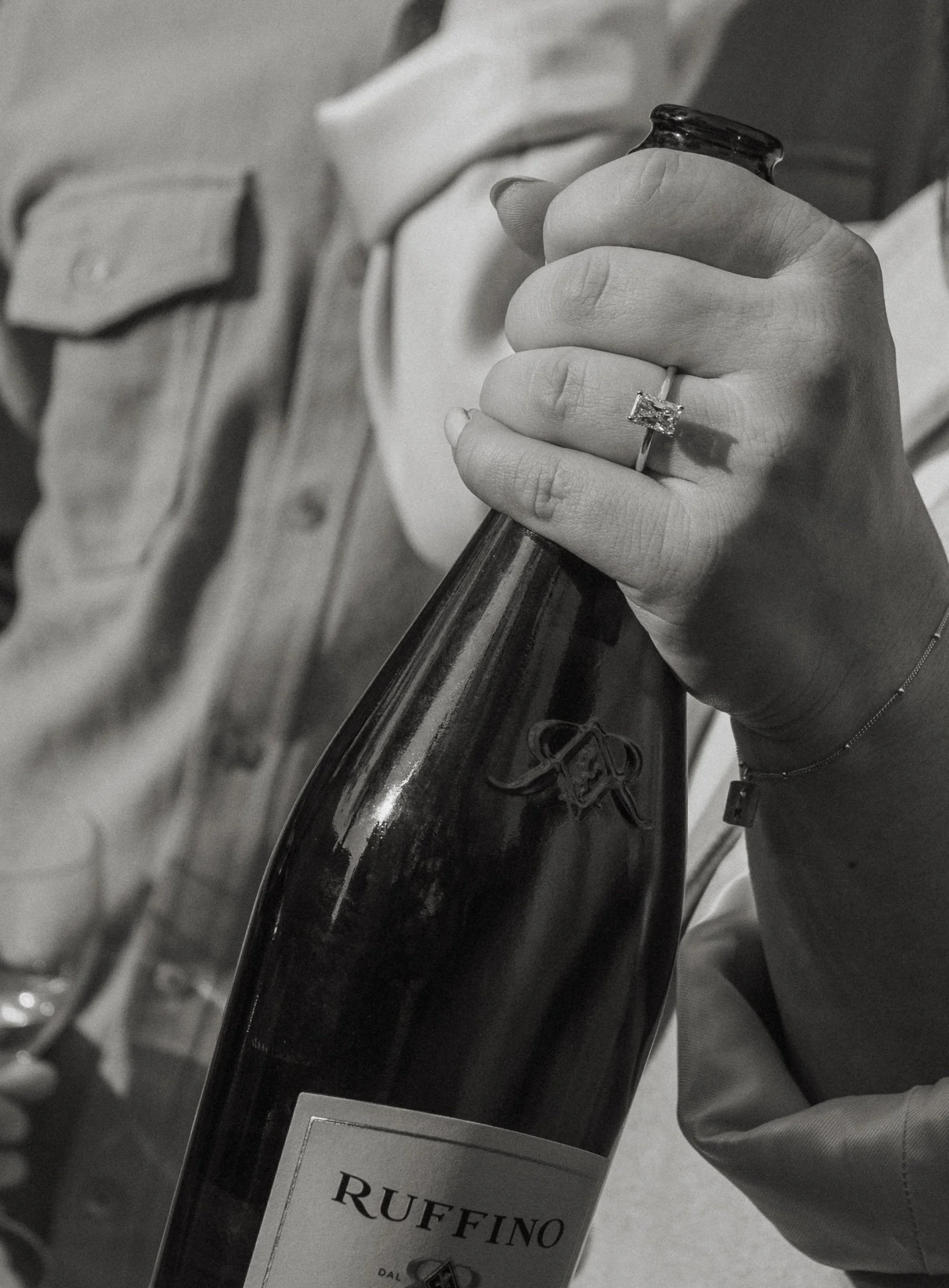 A person holding a bottle of Ruffino wine, wearing a ring with a large square gemstone and a bracelet, with part of their face not visible in the photo.