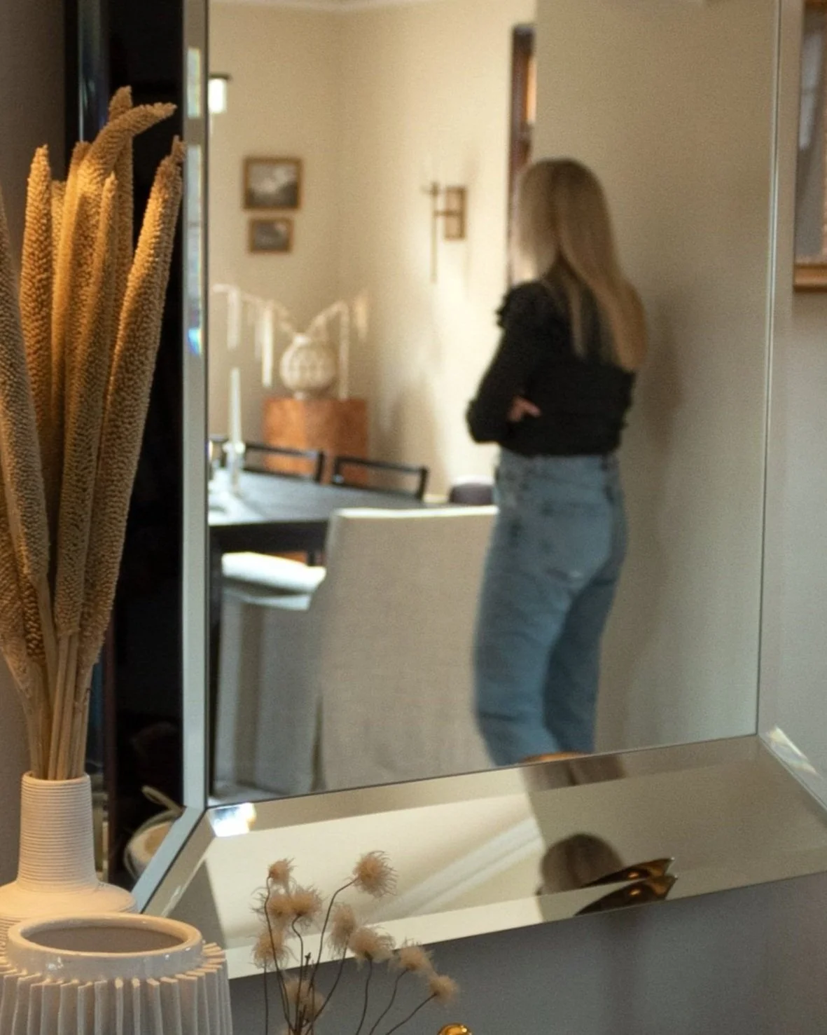 A woman with blonde hair standing in front of a mirror with her arms crossed, in a furnished living or dining area with wall art and a table set with candles in the background.