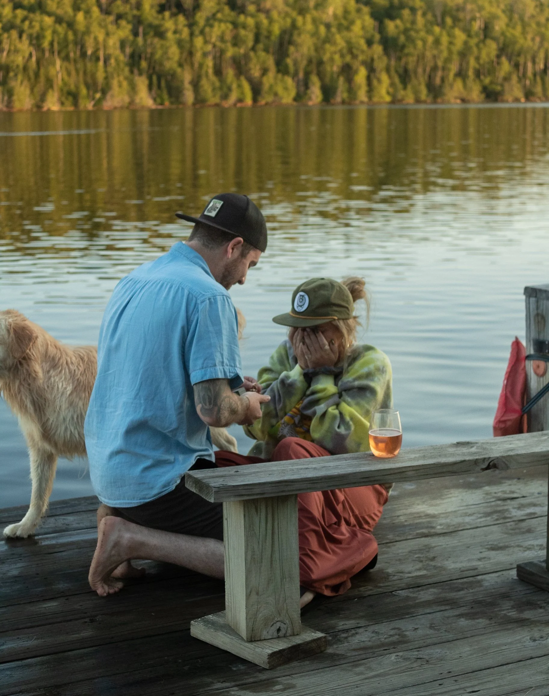A man kneeling on a wooden dock, showing something on his phone to a woman sitting with her hands on her face, near a lake with trees in the background. There is a golden retriever dog and a glass of rosé wine on the dock.