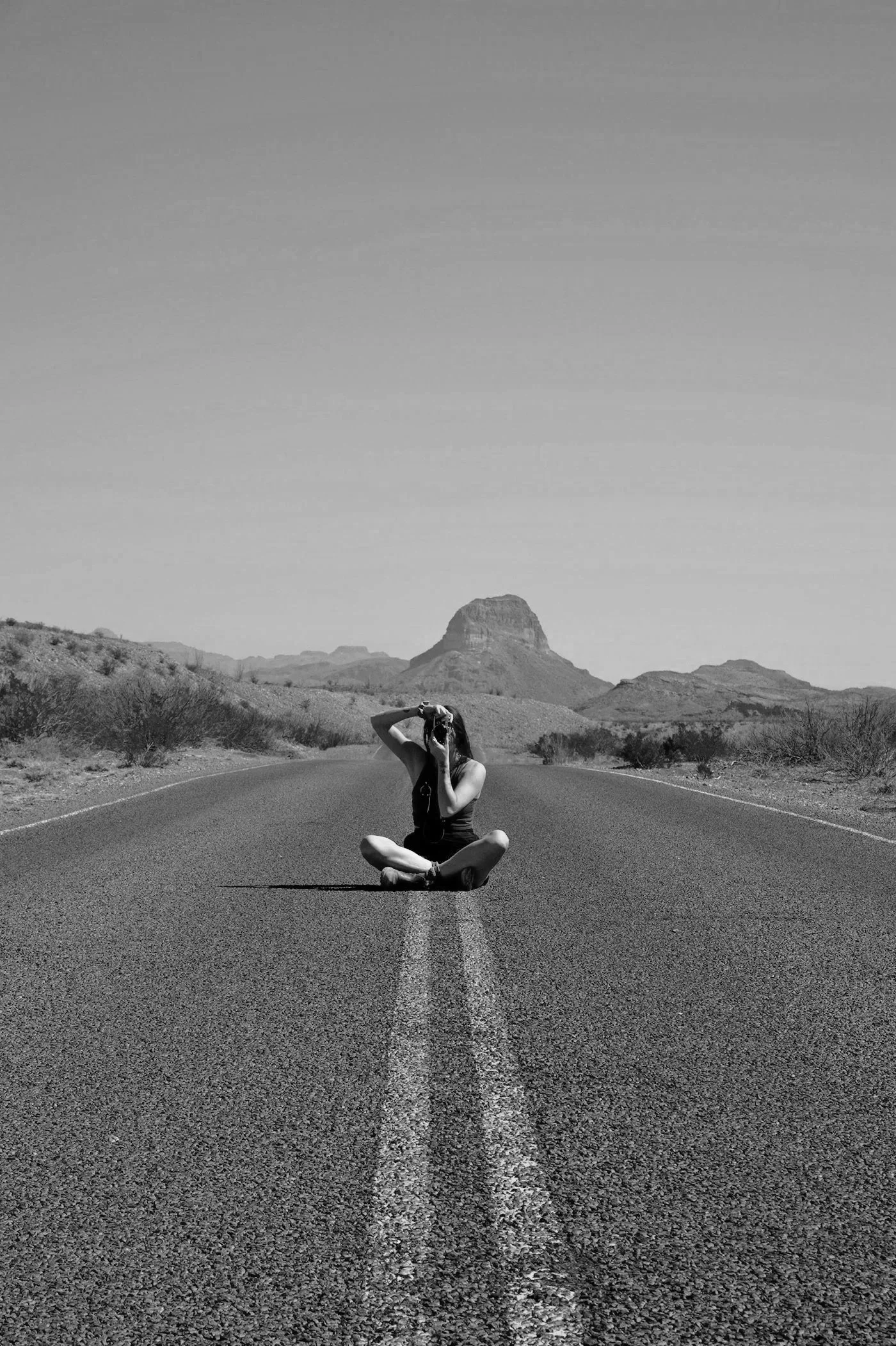 A black-and-white photo of a woman sitting cross-legged in the middle of a deserted open road, taking a photo with a camera. The landscape features desert terrain with a mountain in the background in big bend national park, texas