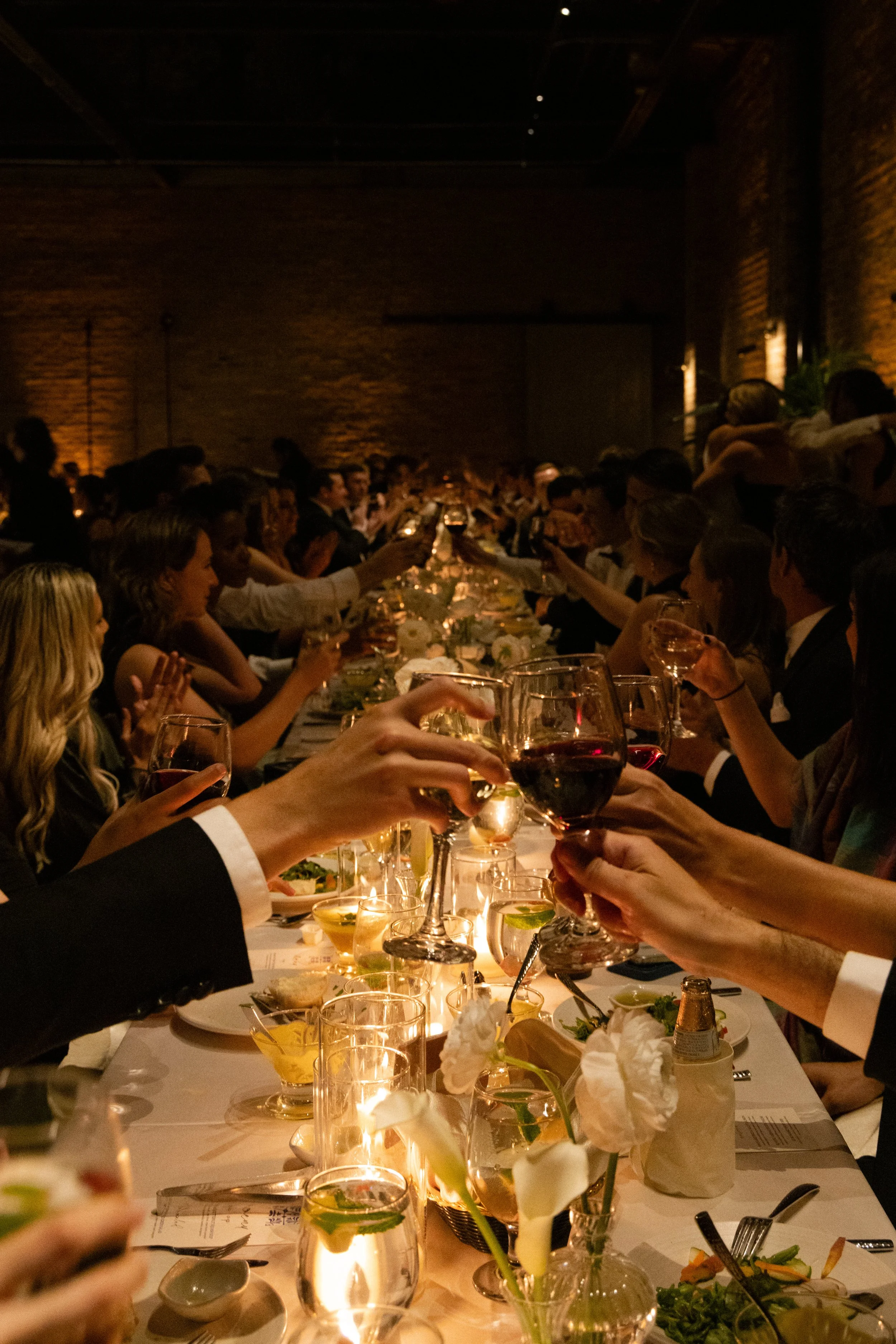 A long banquet table filled with people in formal attire raising their glasses for a toast in a dimly lit, rustic-style event space with brick walls.