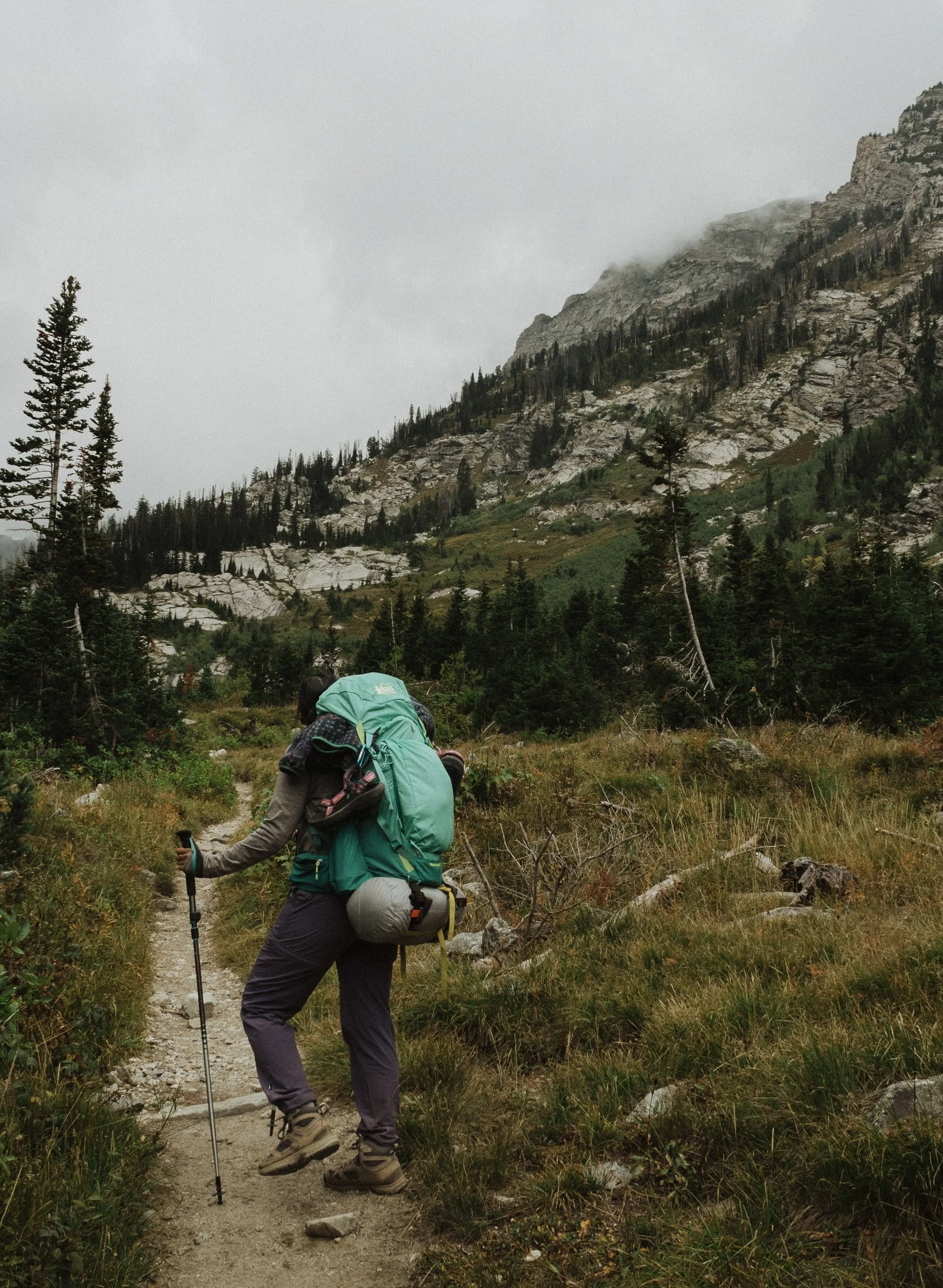 Hiker adjusting a backpack while walking on a mountain trail surrounded by trees and rocky hills under an overcast sky.
