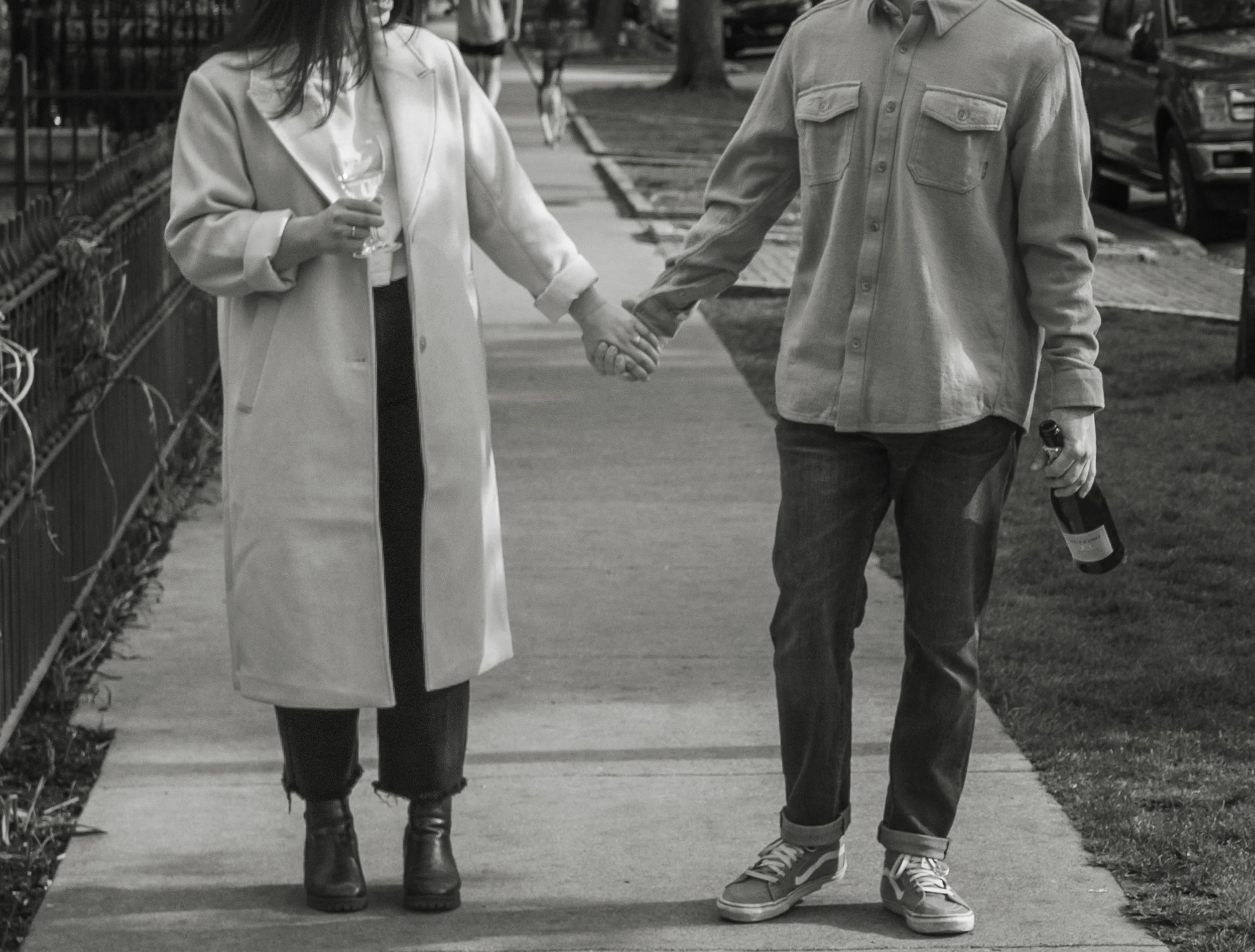 A man and a woman holding hands while walking on a city sidewalk. The woman is holding a glass of wine, the man is holding a bottle. Both are dressed casually, the woman in a long coat and boots, the man in a button-up shirt and sneakers.