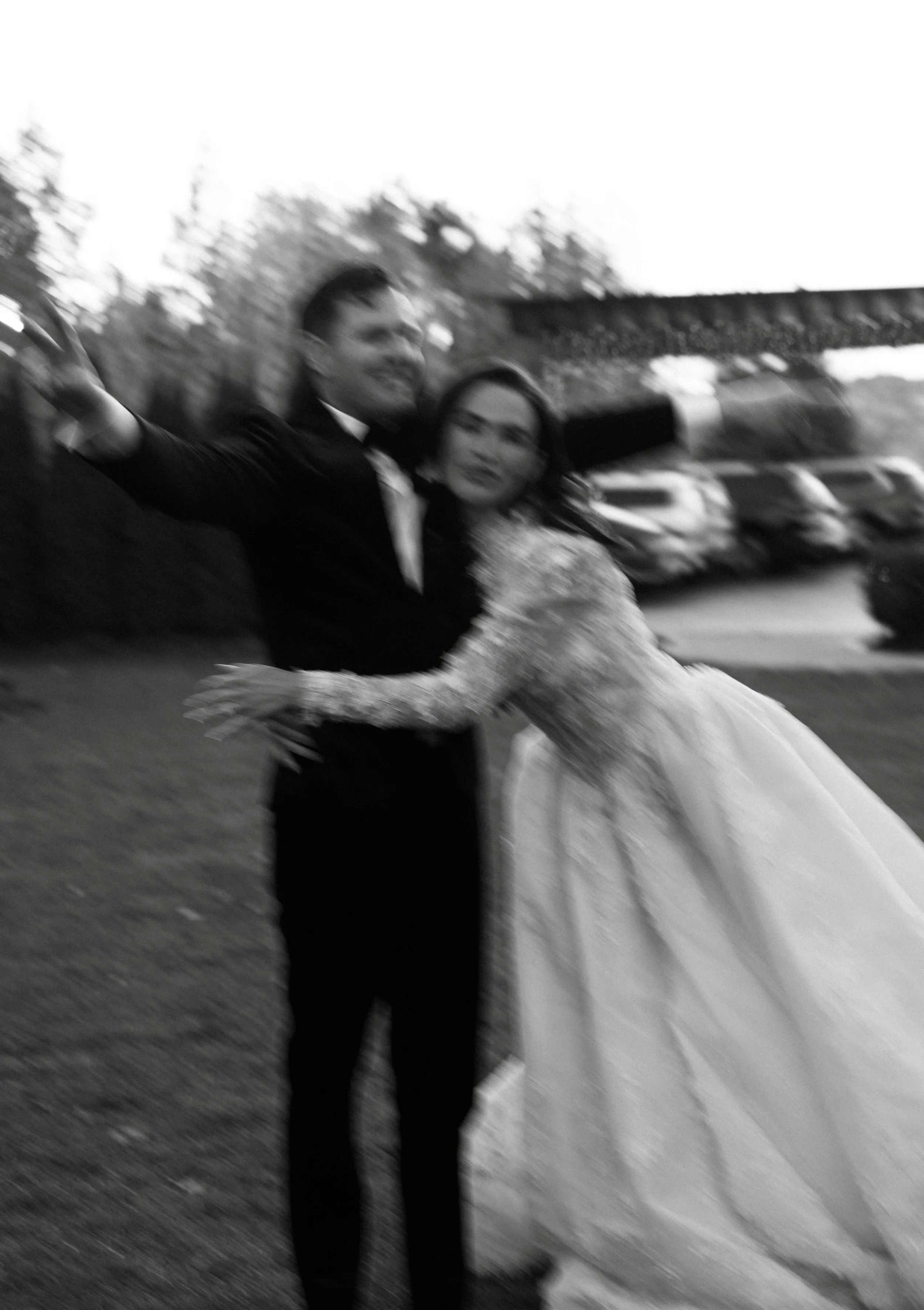 a motion blurred black-and-white photo of a couple on their wedding day, the man in a tuxedo and the woman in a wedding dress, embracing each other outdoors.