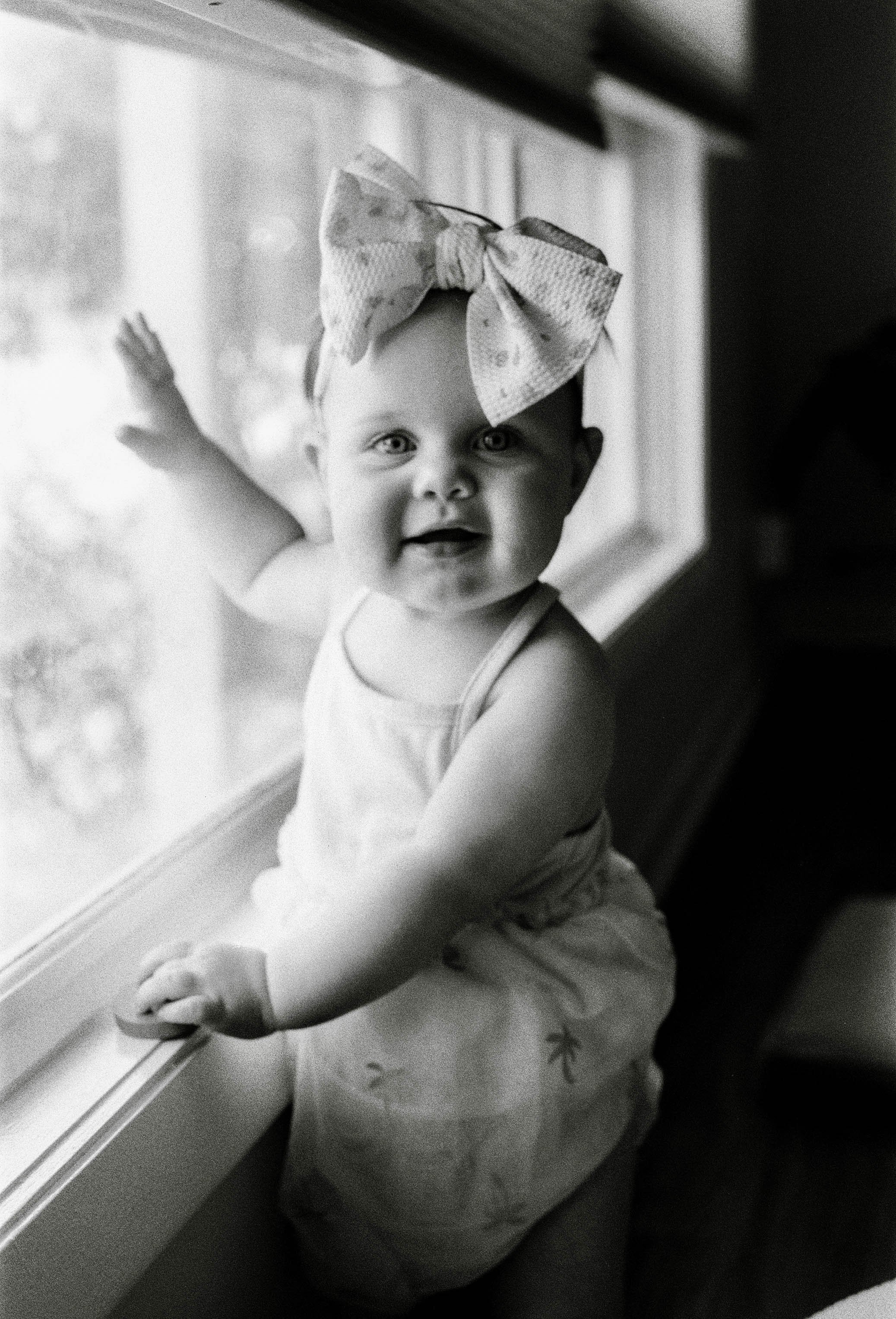 A smiling baby girl with a large bow on her head, leaning on a windowsill and looking at the camera.