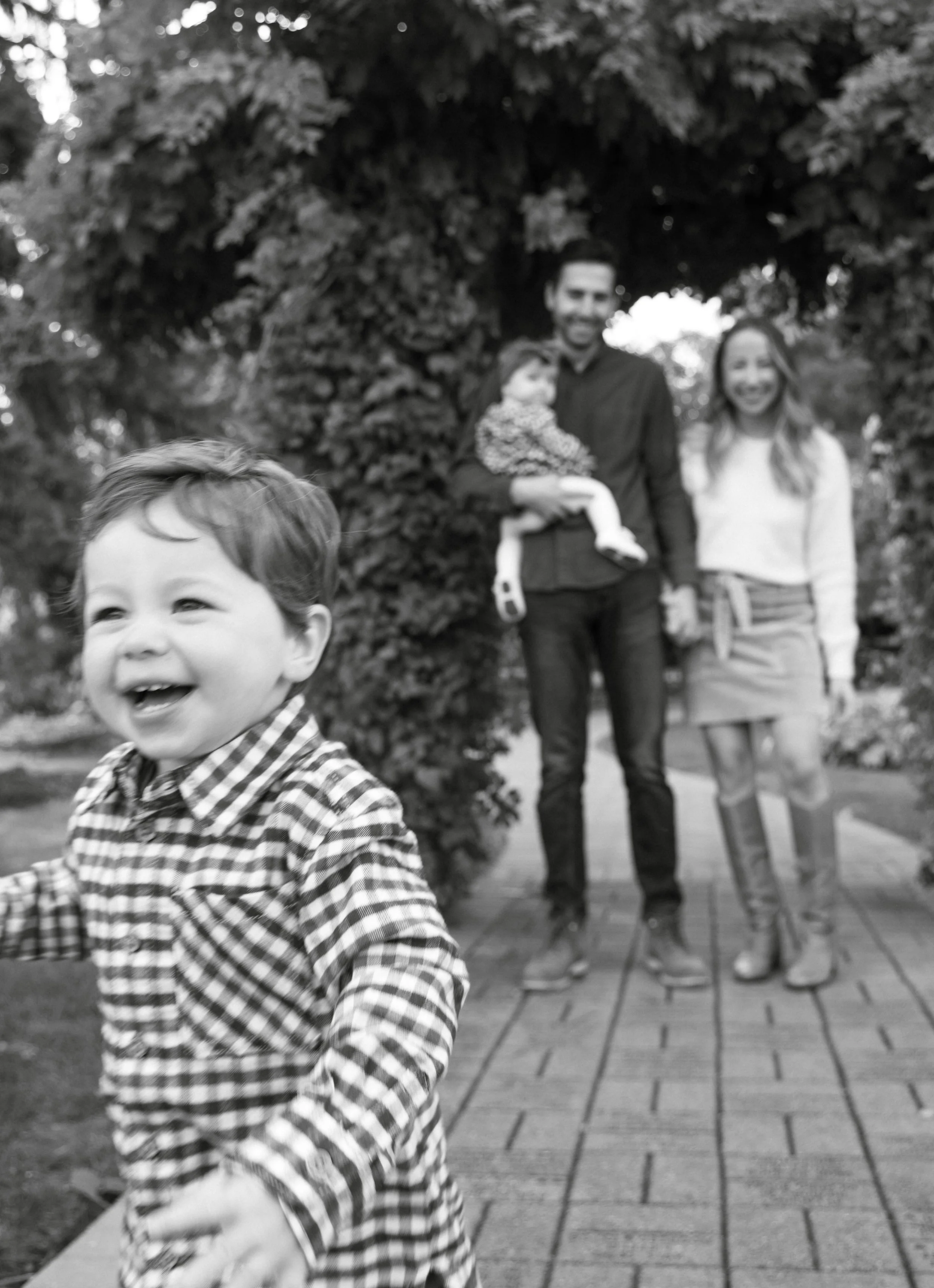 A young boy with a checkered shirt smiling and running outdoors, with a family of three in the background near a large tree and some bushes.