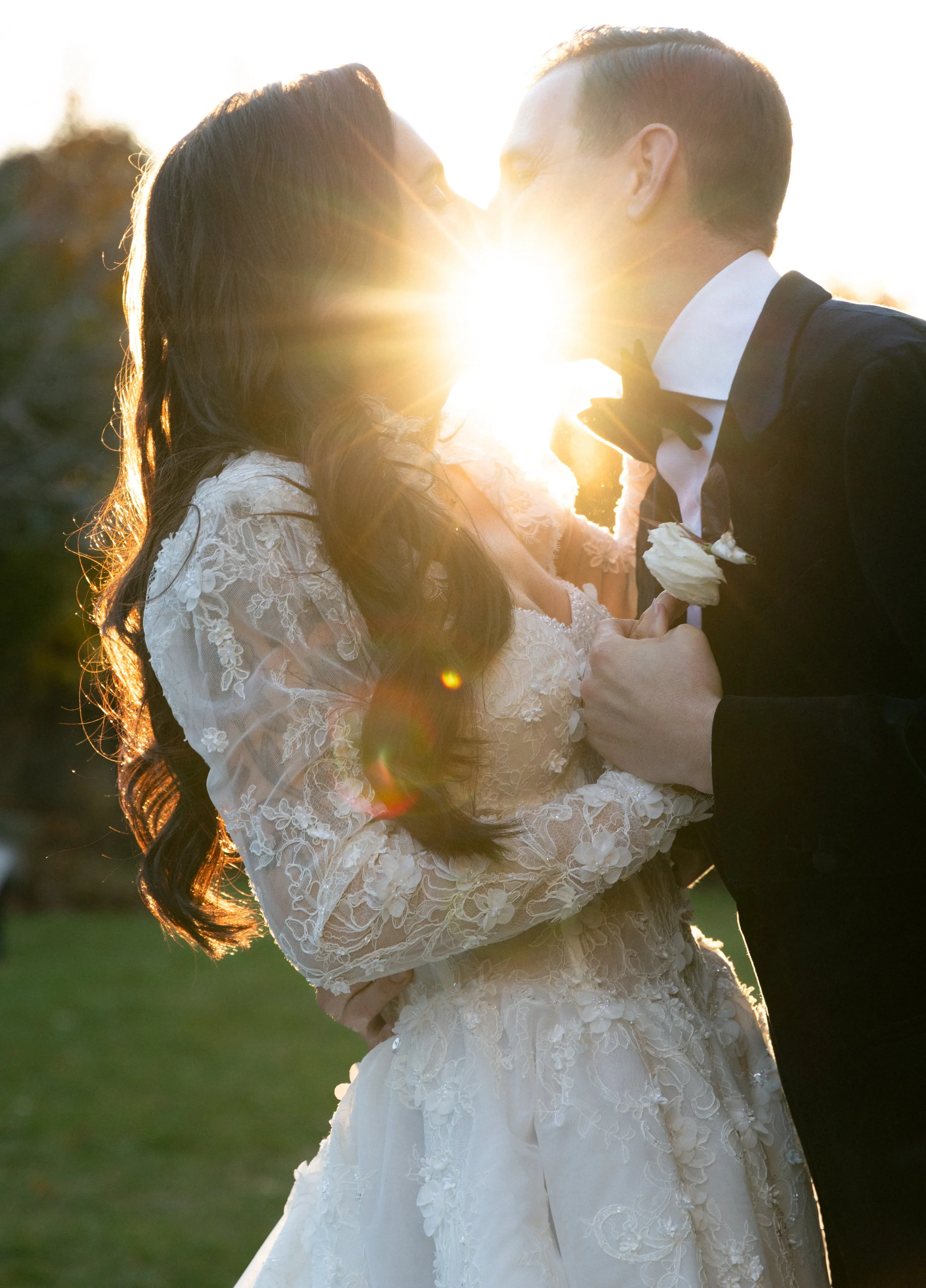 A newlywed couple sharing a kiss outdoors at sunset, with the sun shining between their faces. The bride wears a lace wedding gown and the groom in a tuxedo with a bow tie, holding a flower in his hand.