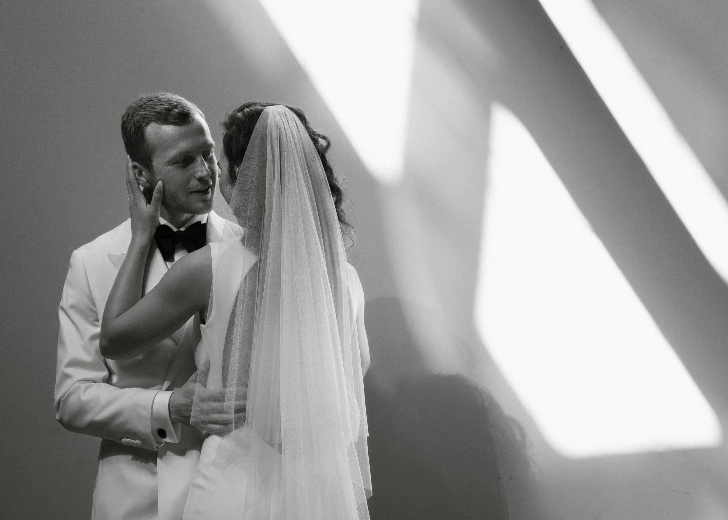 A black and white photo of a bride and groom sharing an intimate moment, with the groom wearing a white tuxedo and the bride in a white wedding dress and veil, standing near a wall with abstract shadows.