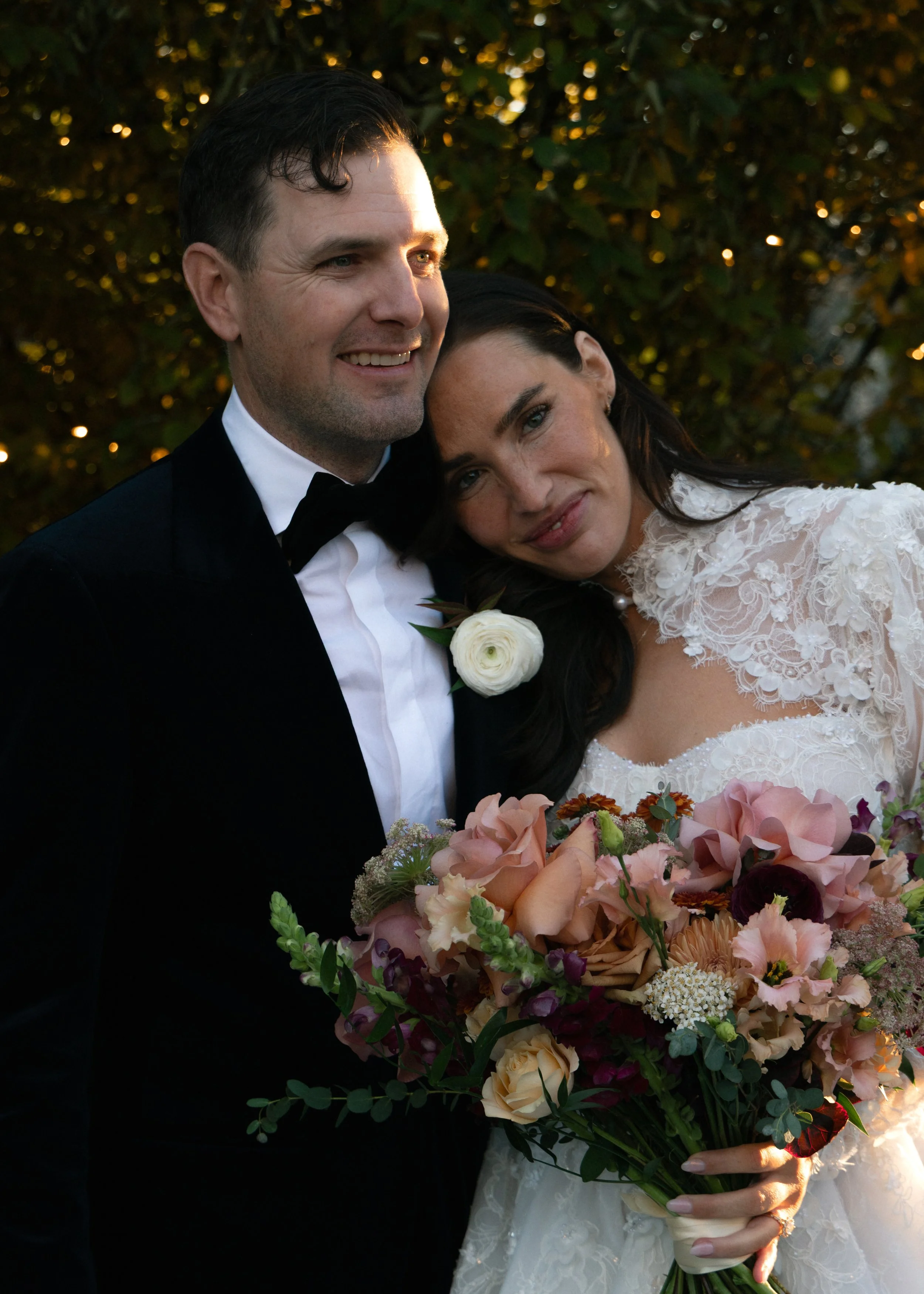 A bride and groom on their wedding day, standing close together outdoors with a leafy background, smiling and holding a large bouquet of pink, peach, and purple flowers.