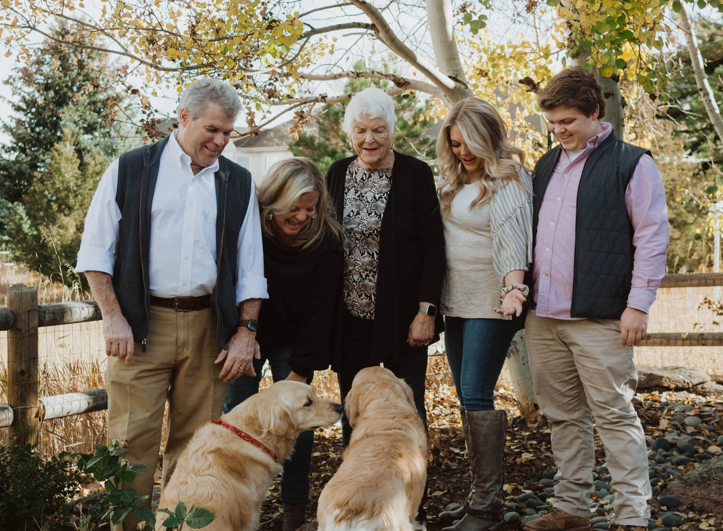 Six people, three women and three men, stand outdoors in autumn, smiling and engaging with two golden retrievers. The group is near a wooden fence and trees with yellow leaves.