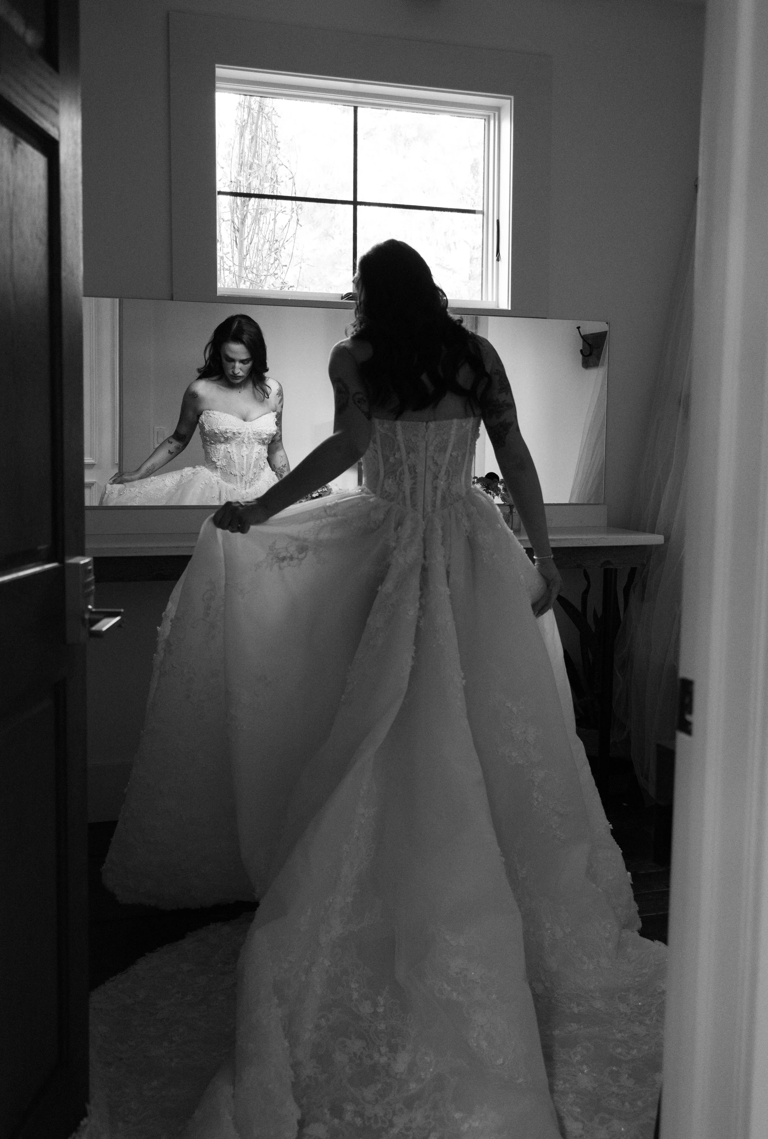 A woman in a wedding dress standing in front of a mirror, adjusting her gown, with a window in the background.
