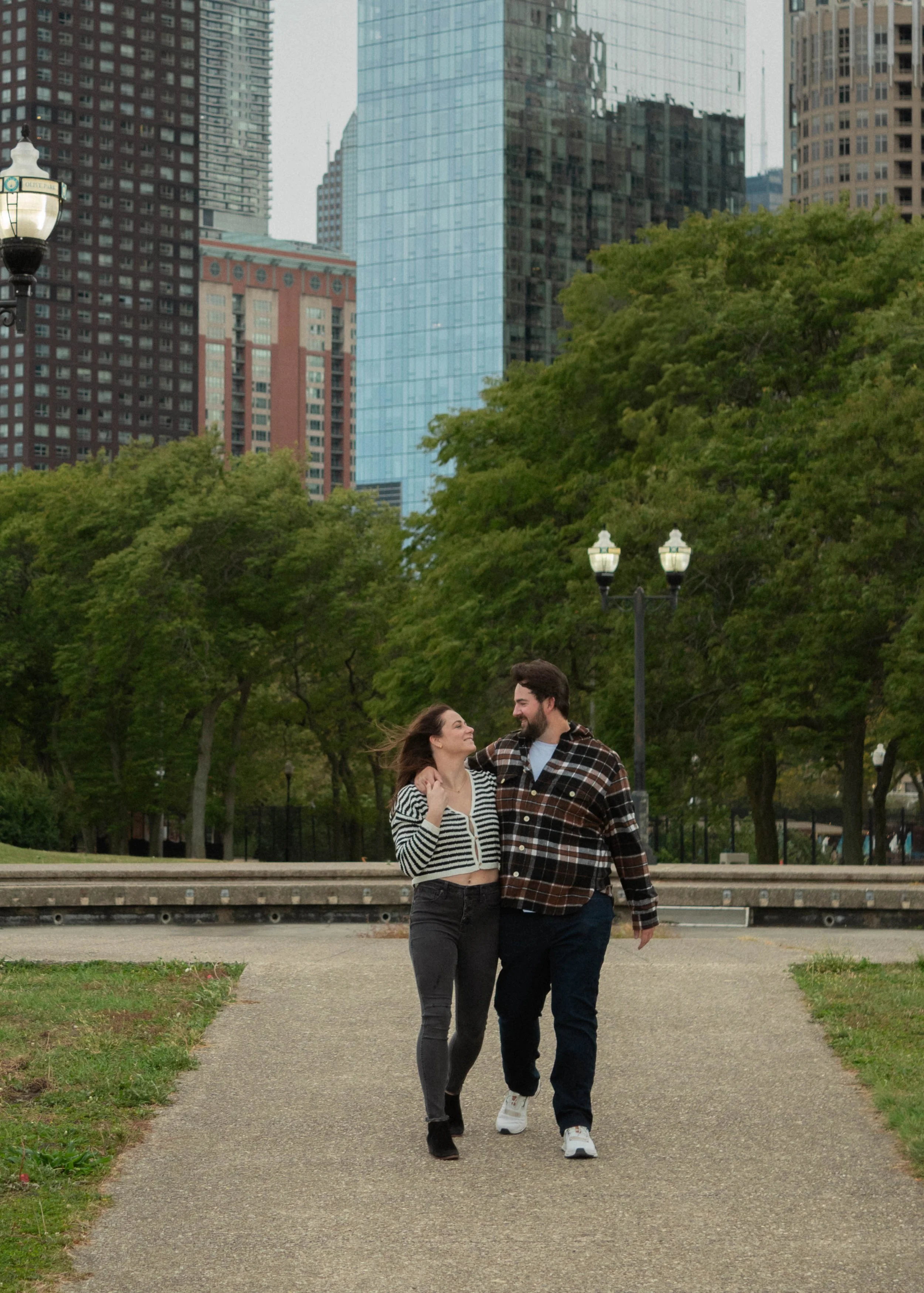 A smiling couple walks together in a park with trees and city buildings in the background.