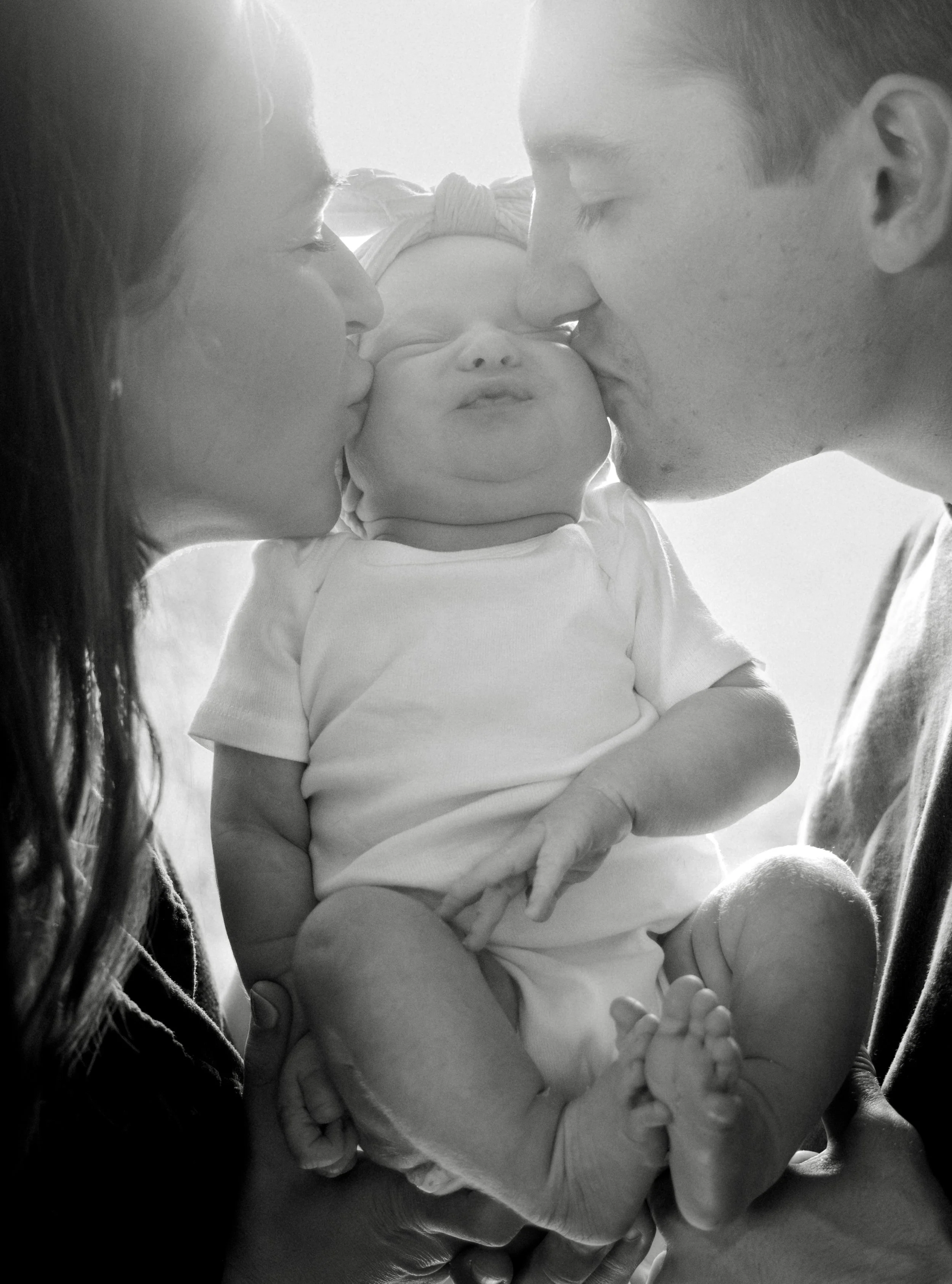 A black and white photo of a newborn baby being kissed twice by her mother and father on each cheek, with the baby wearing a headband and lying on an adult's hand, in an emotional family moment.