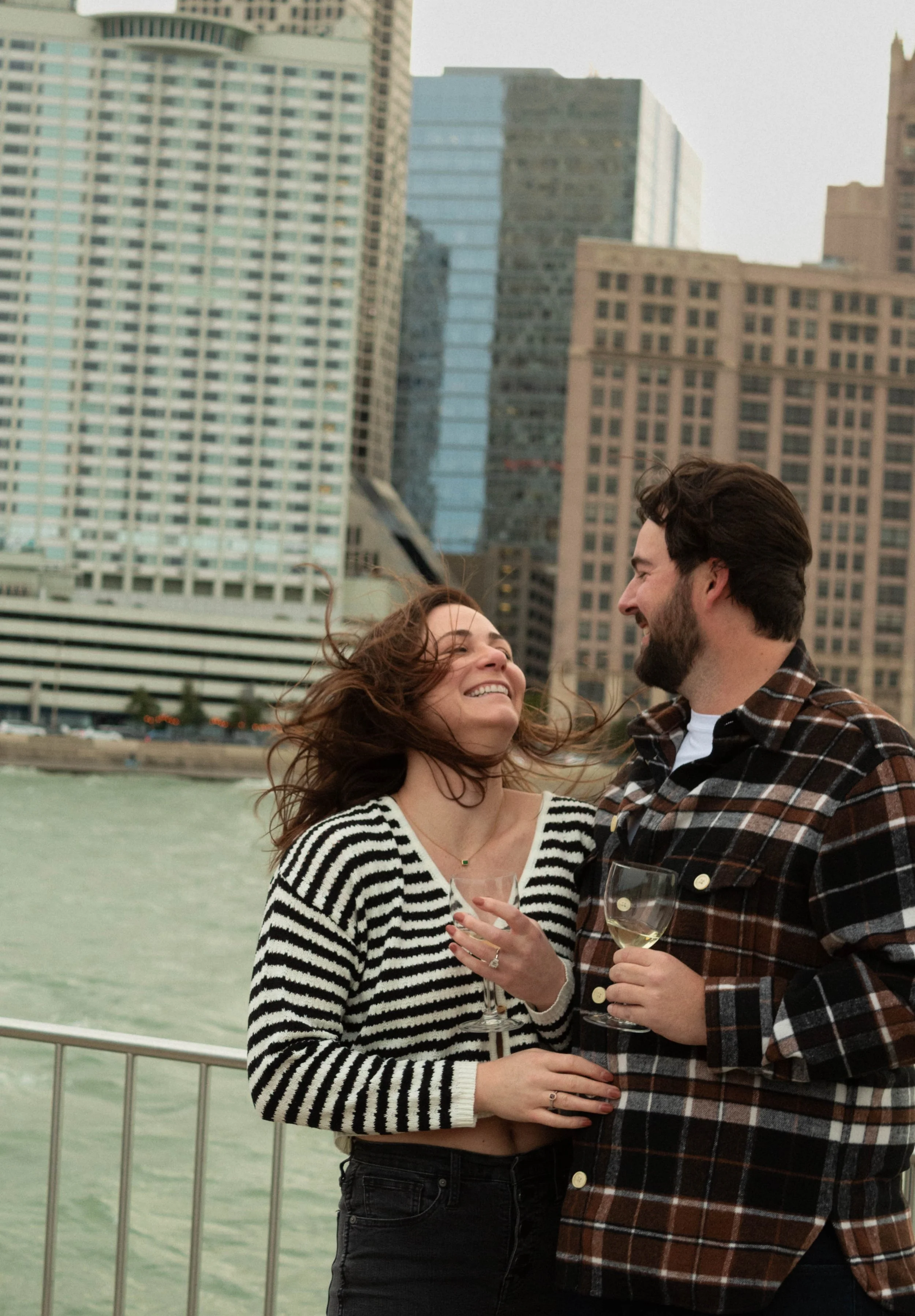 A smiling woman and a man holding wine glasses are outdoors near water with tall buildings in the background.