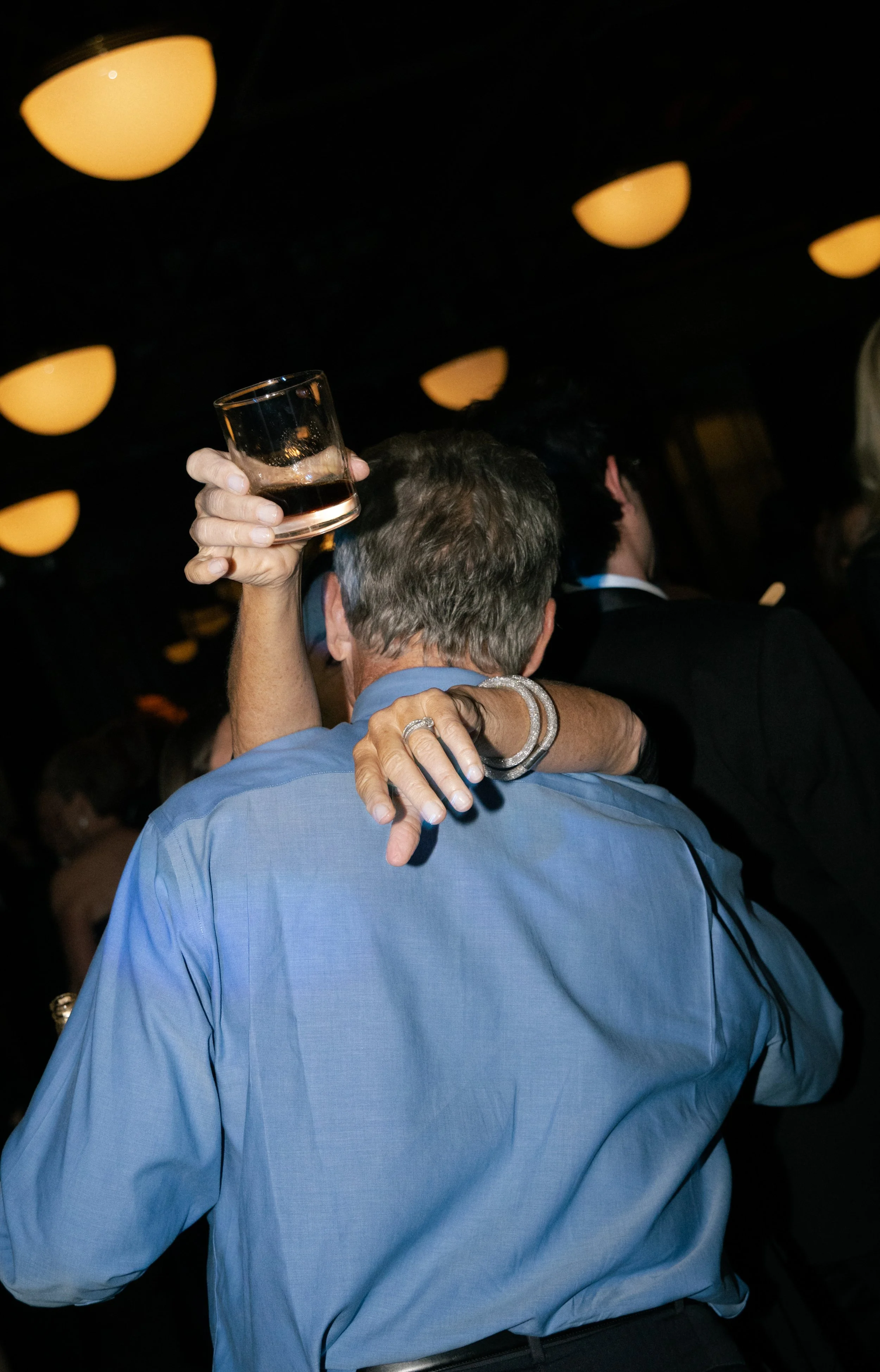 A woman hugging a man at a party or celebration, with the woman holding a glass of dark beverage, in a dimly lit room with orange ceiling lights.