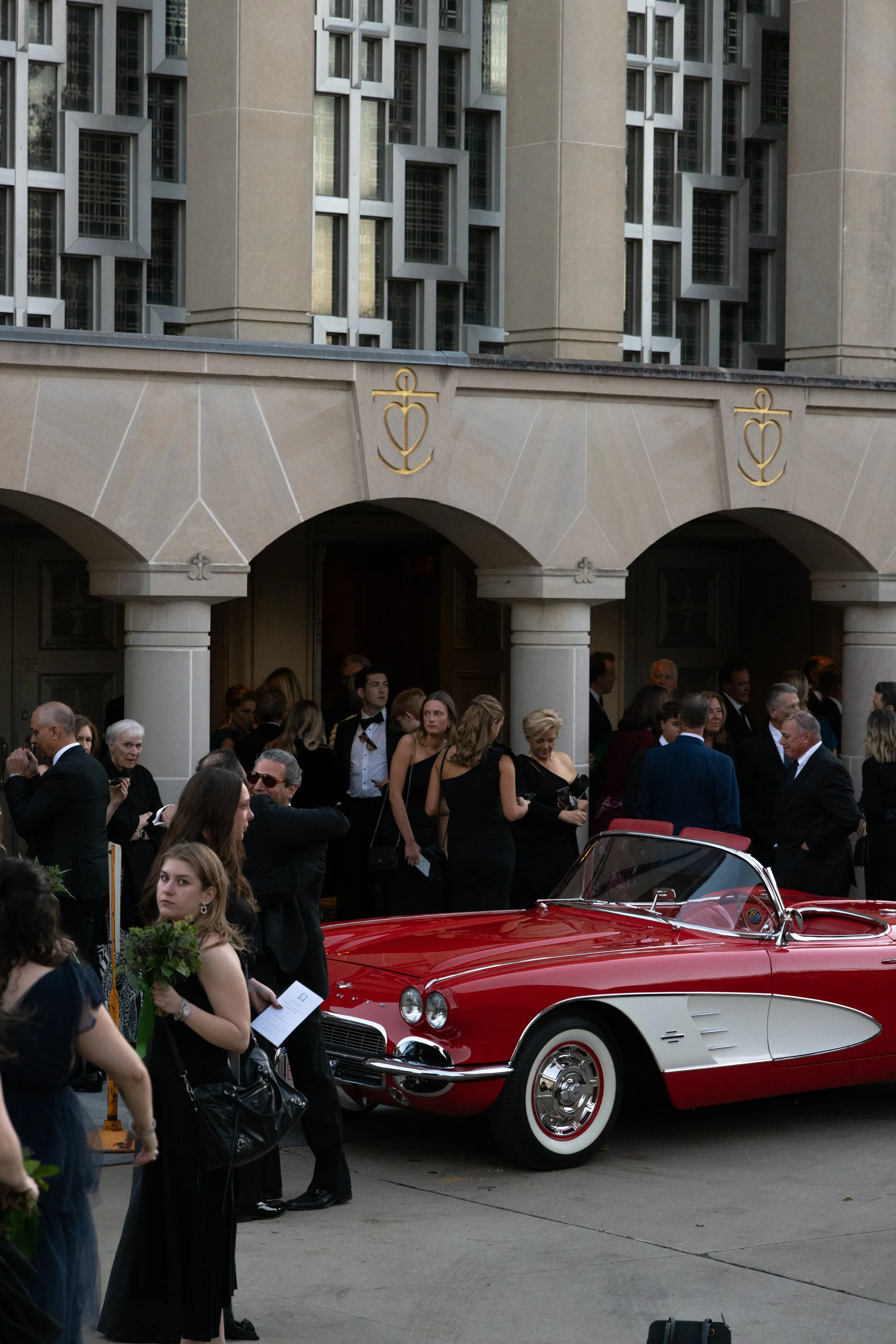 People dressed in formal attire gather outside a building, with a vintage red and white convertible car parked in front.