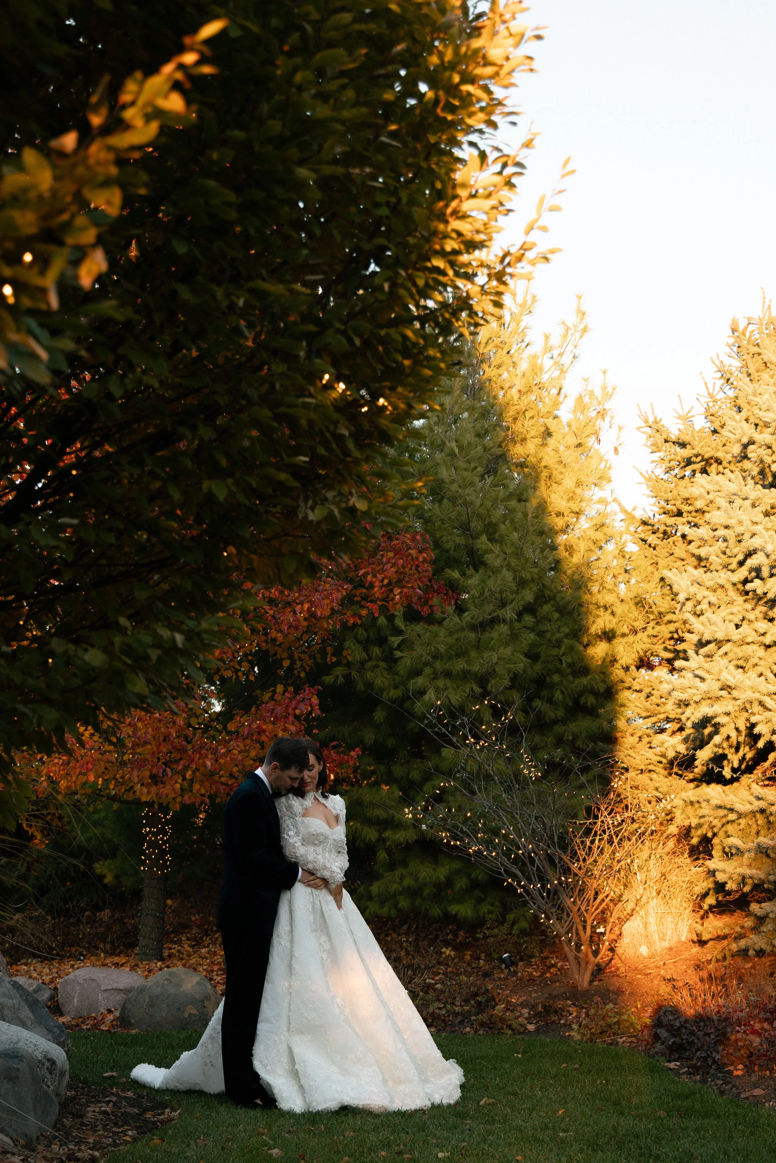 A bride and groom standing close together outdoors during sunset, surrounded by colorful trees and outdoor lighting, with rocks and grass underneath.