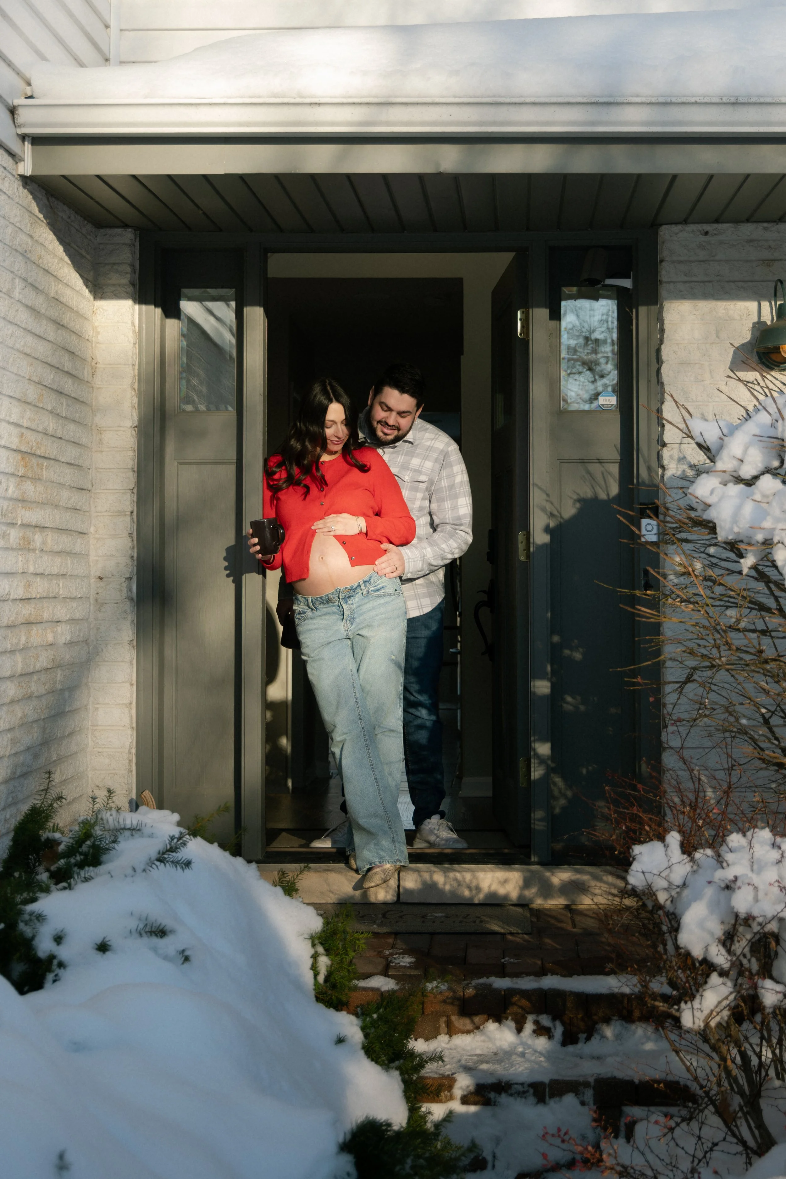 A couple standing at the doorway of a house with snow outside. The woman, who appears pregnant, is lifting her red sweater to show her belly, and the man is standing behind her, smiling and gently touching her belly.