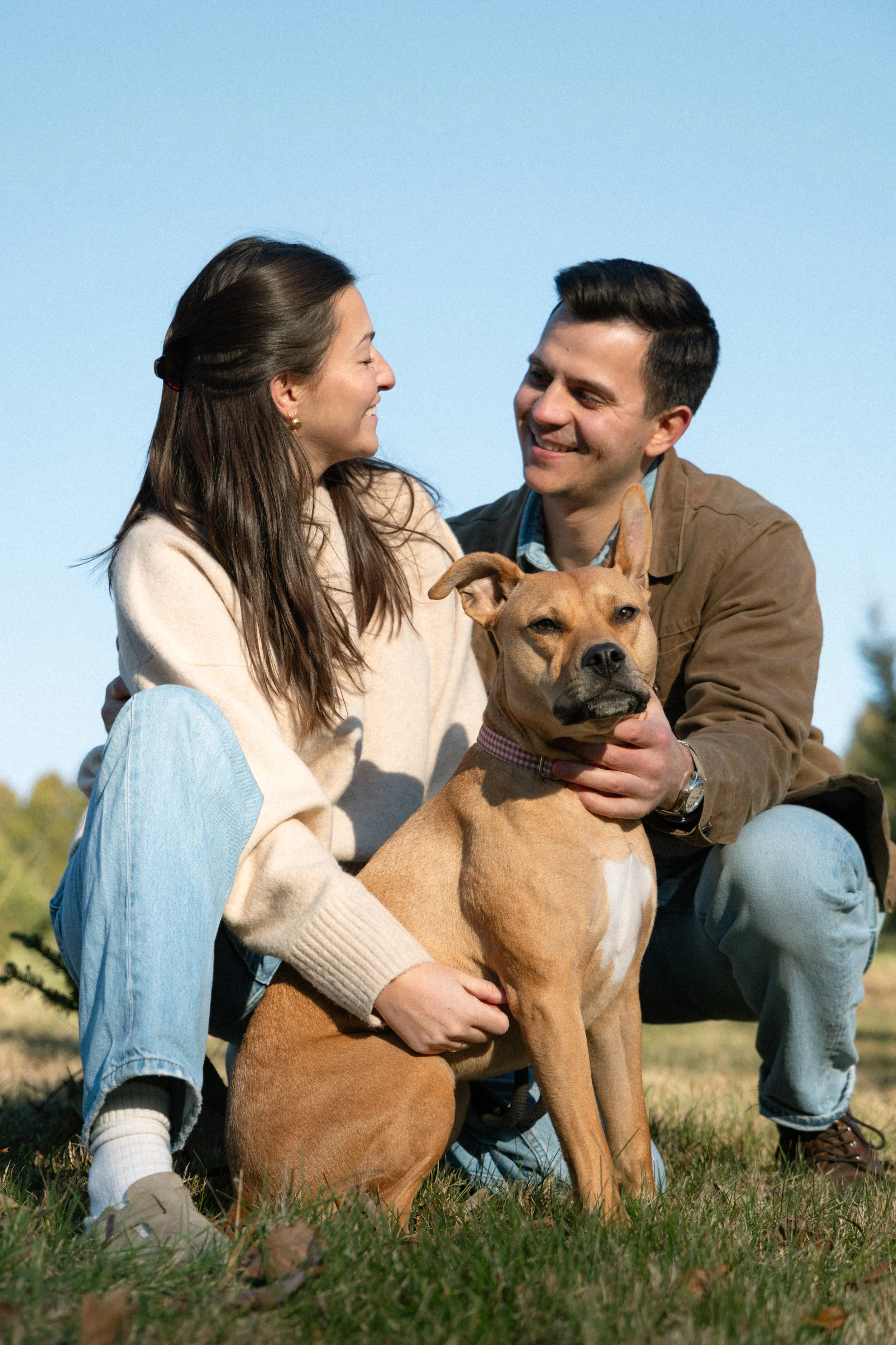A couple with a dog outdoors on a grassy field on a clear day, smiling at each other.