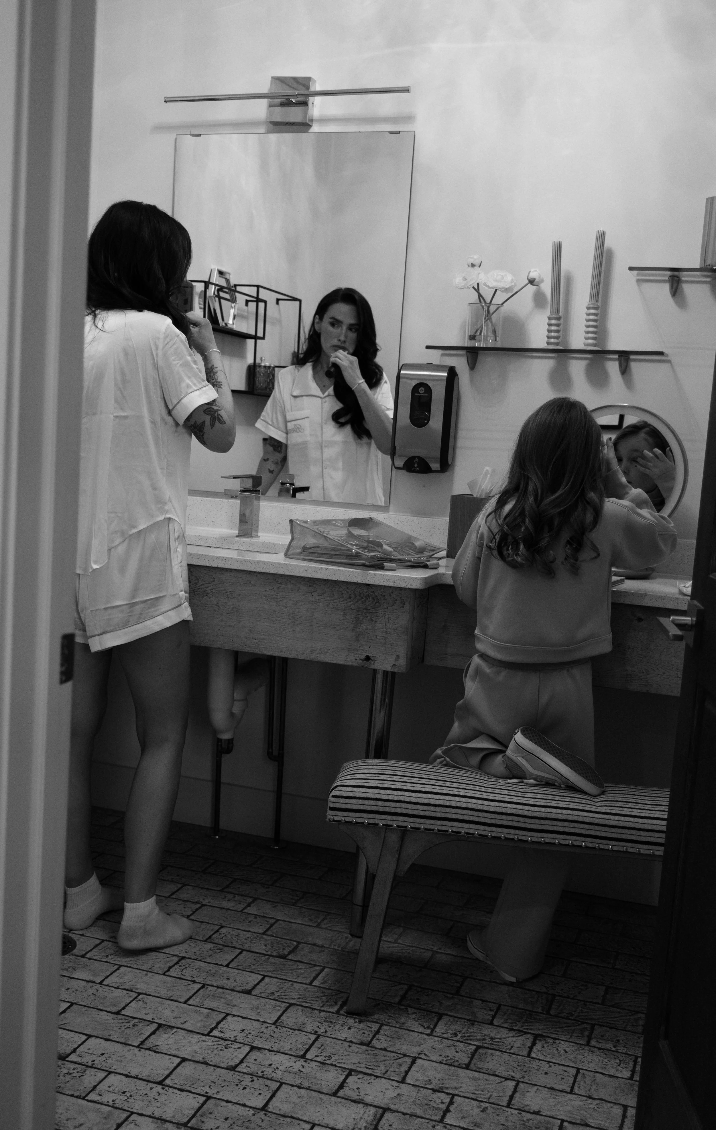 A woman and a young girl are getting ready in a bathroom. The woman is looking at her reflection in the mirror while the girl is applying makeup or skincare at the sink.