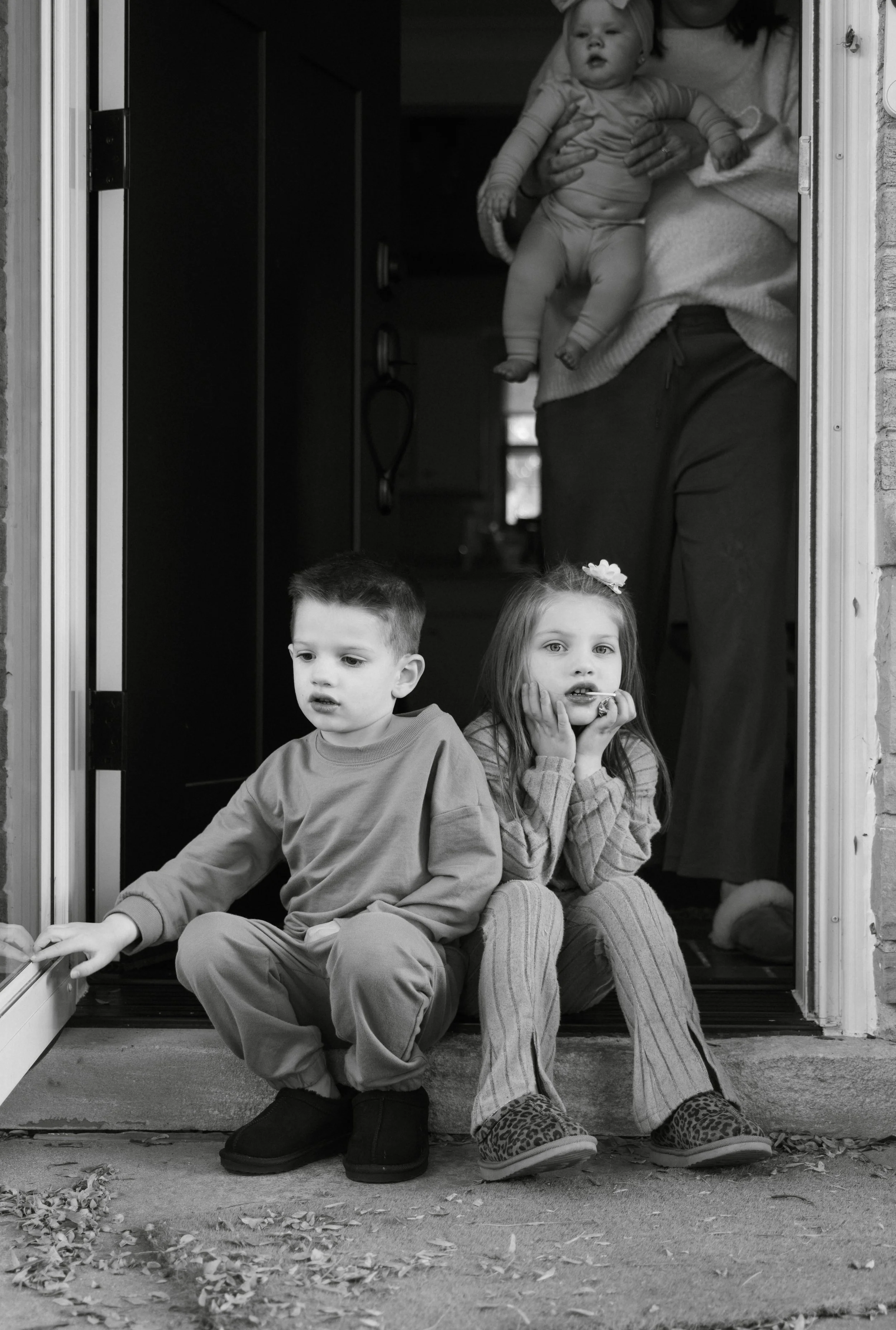 Two children sitting on a porch step, a boy and a girl, with a woman holding a baby standing in the doorway behind them. The children appear concerned or puzzled.