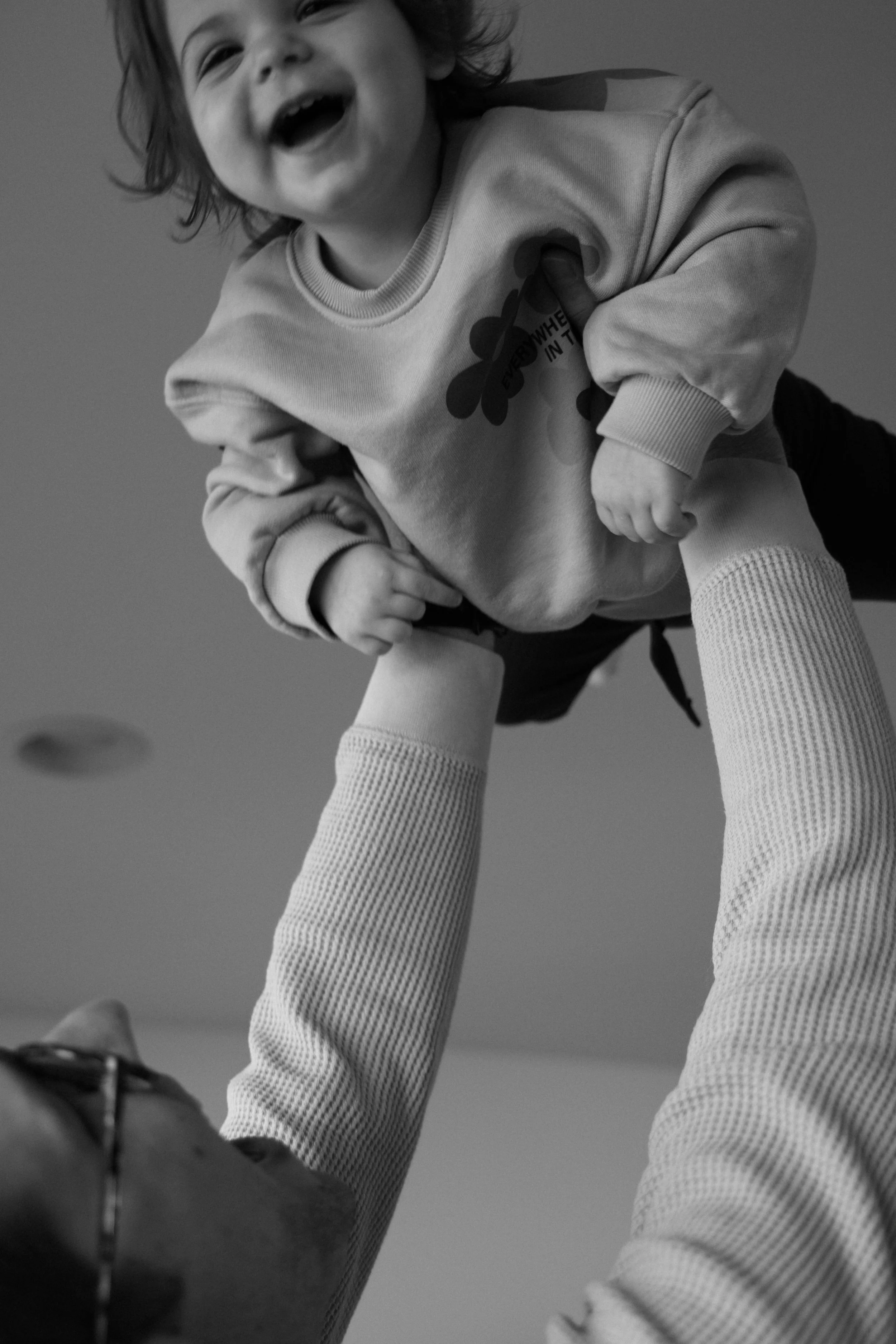 A young child laughing happily while being lifted by an adult, visible only by her arms, in a playful moment indoors.