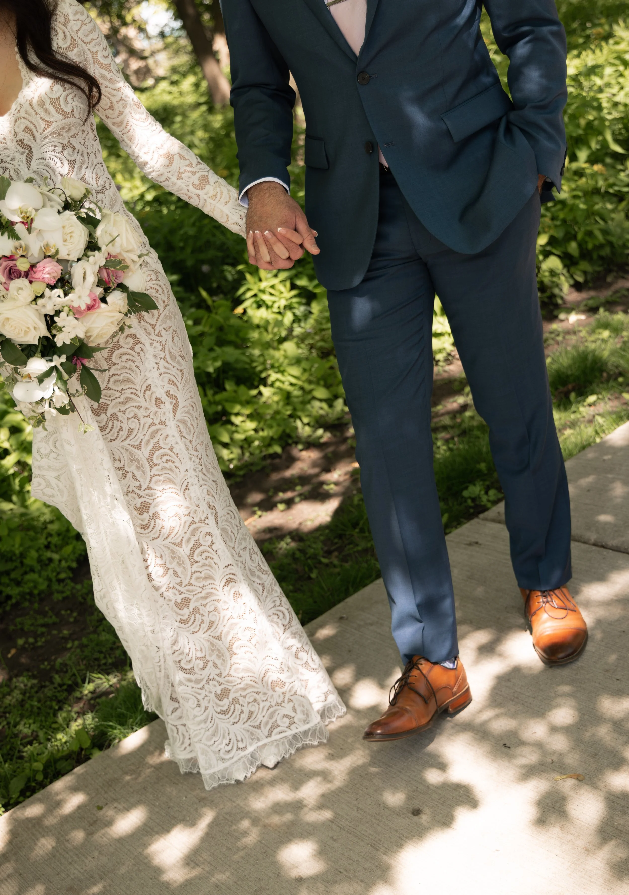 A bride and groom walking hand in hand outdoors on a sidewalk, with the bride in a lace wedding dress holding a bouquet, and the groom in a navy suit with brown shoes.