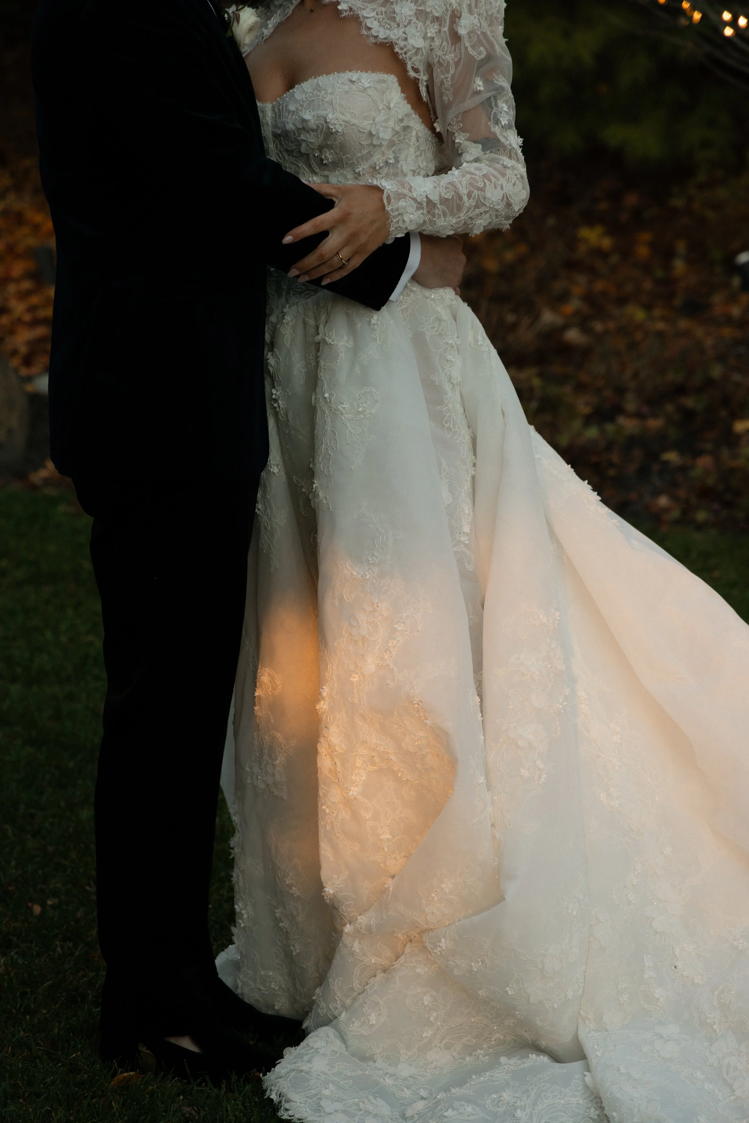 A bride and groom embrace outdoors during sunset, with the bride wearing a lace wedding gown and the groom in a black tuxedo.