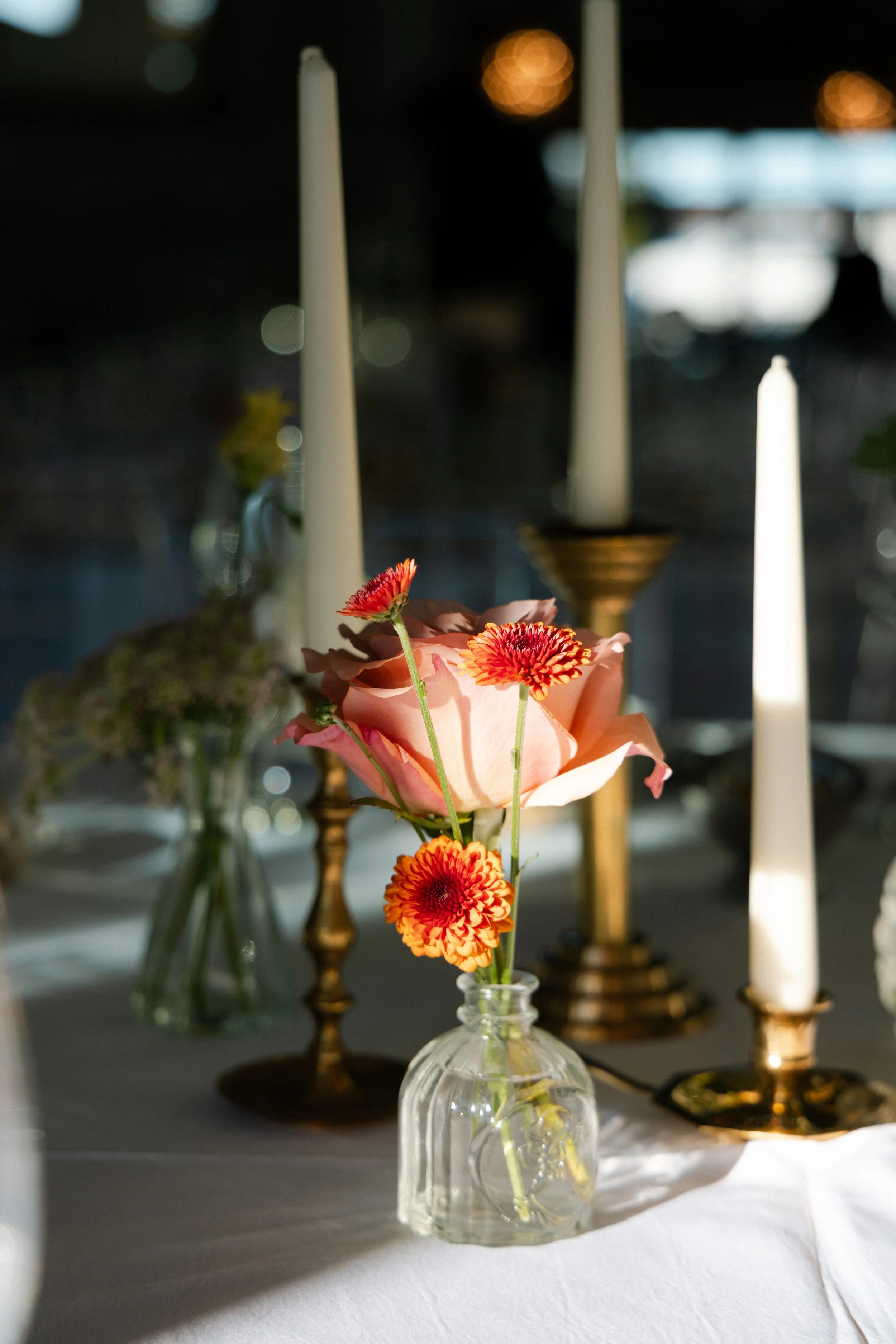 A small glass vase holding pink roses and orange flowers, on a table with gold candlesticks and white candles.