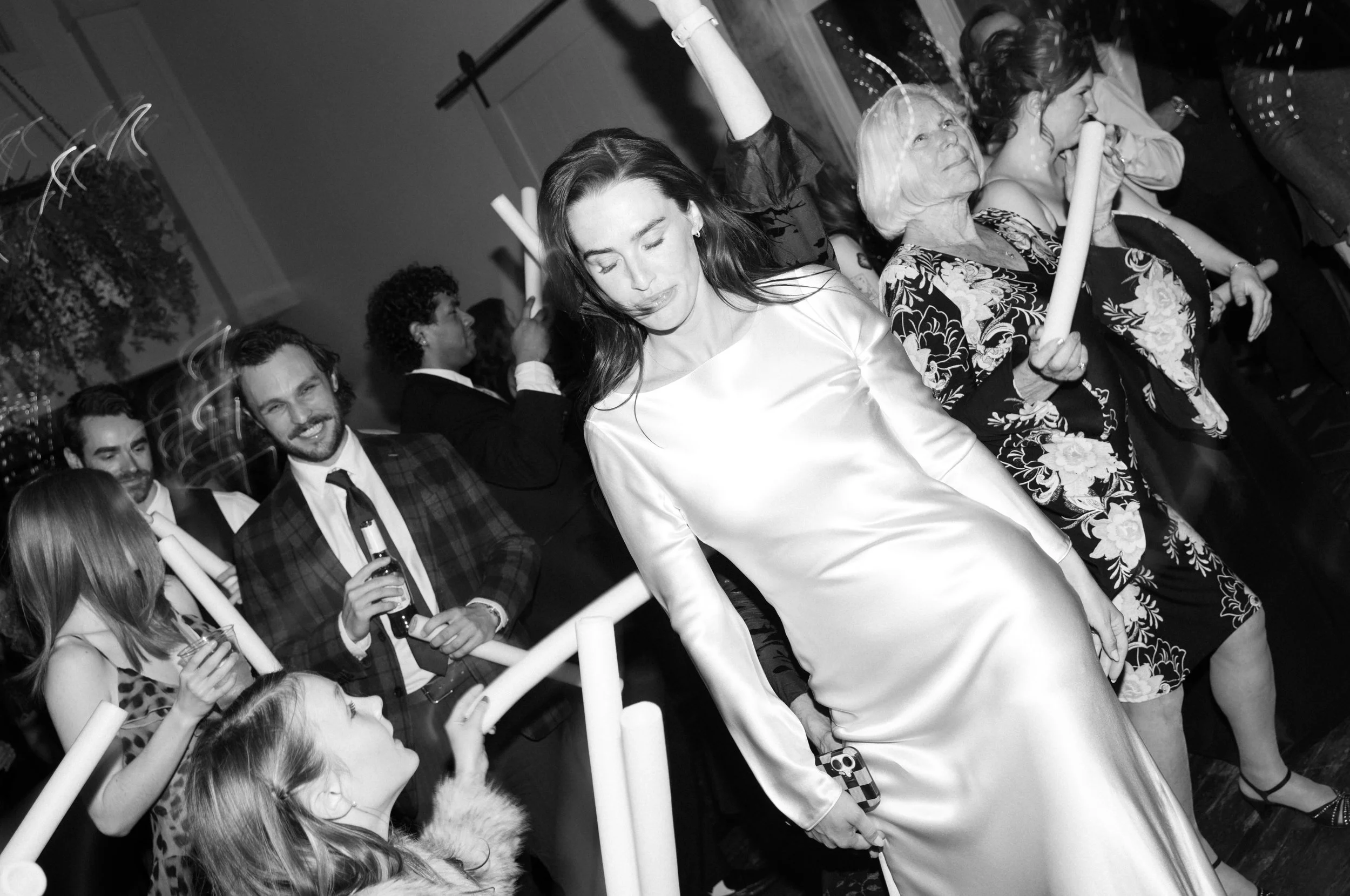 People dancing and celebrating at a party or wedding, with a woman in a satin gown in focus, surrounded by others holding candles and drinking.