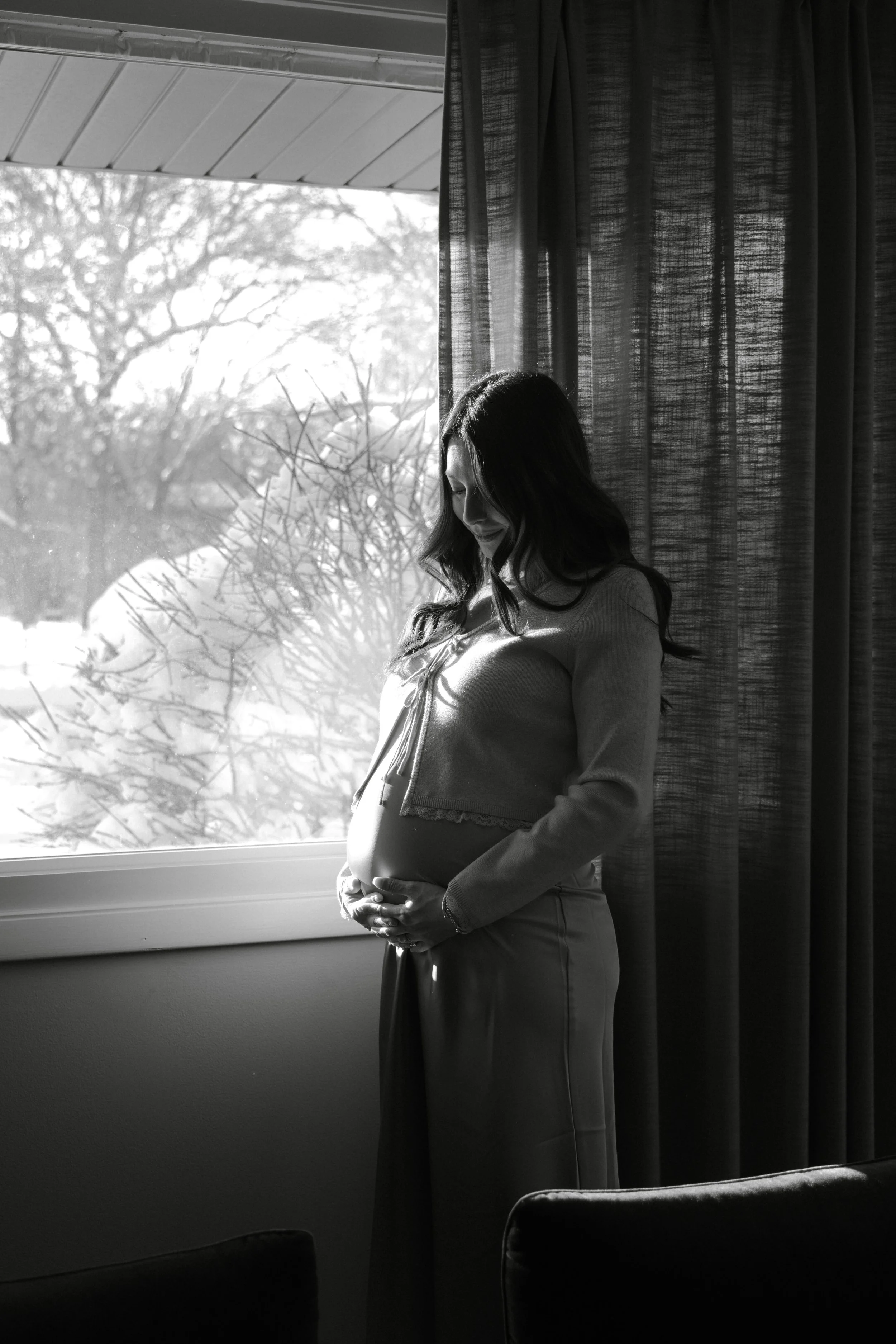 Black and white photo of a pregnant woman standing by a window, smiling and holding her belly, with trees outside.