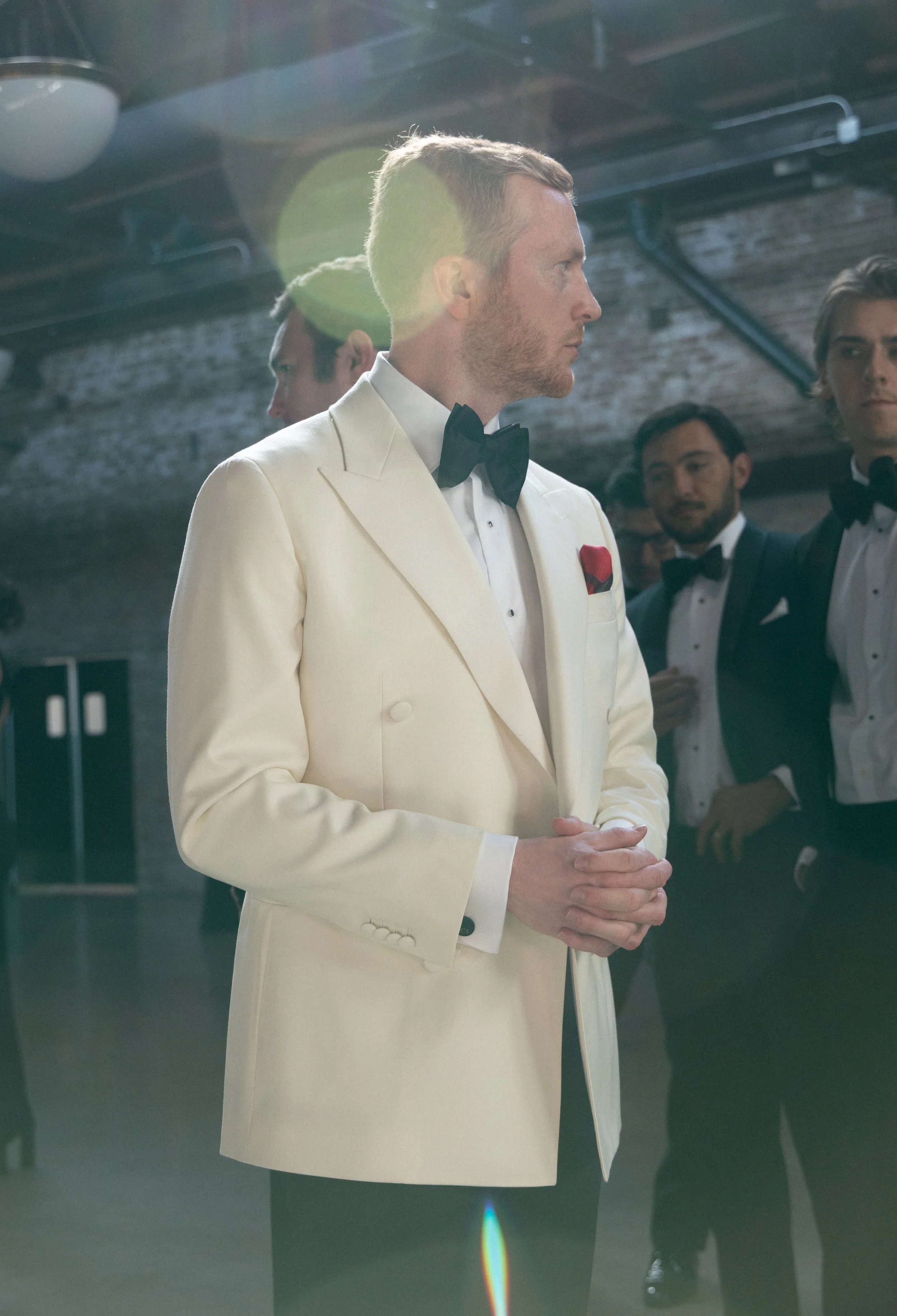 A man in a cream-colored tuxedo with a black bow tie and a red pocket square, standing with hands clasped, at a formal event with others in tuxedos in the background.