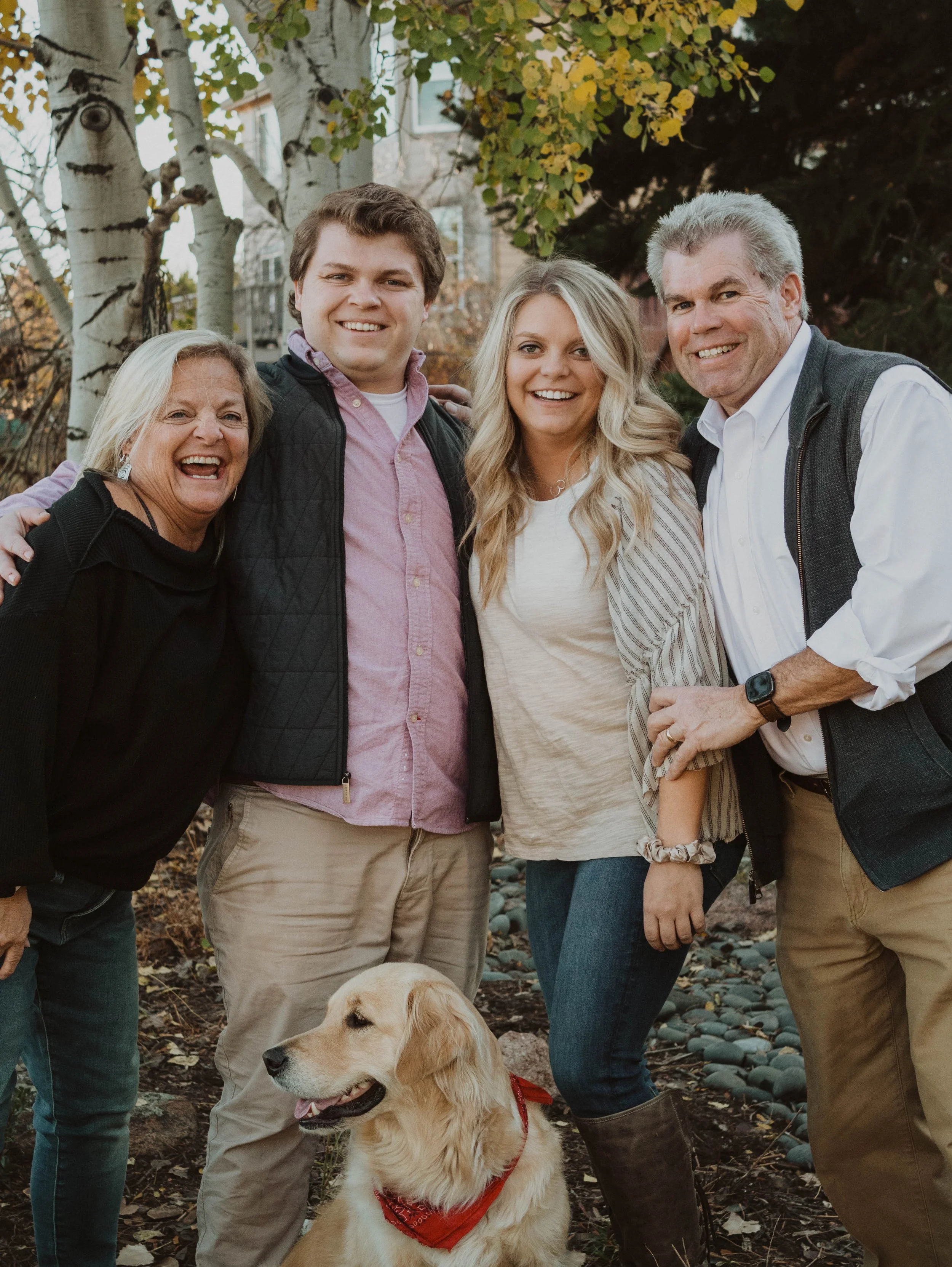 A smiling family of five, including a Golden Retriever dog with a red bandana, standing outdoors in a park with trees and a building in the background.