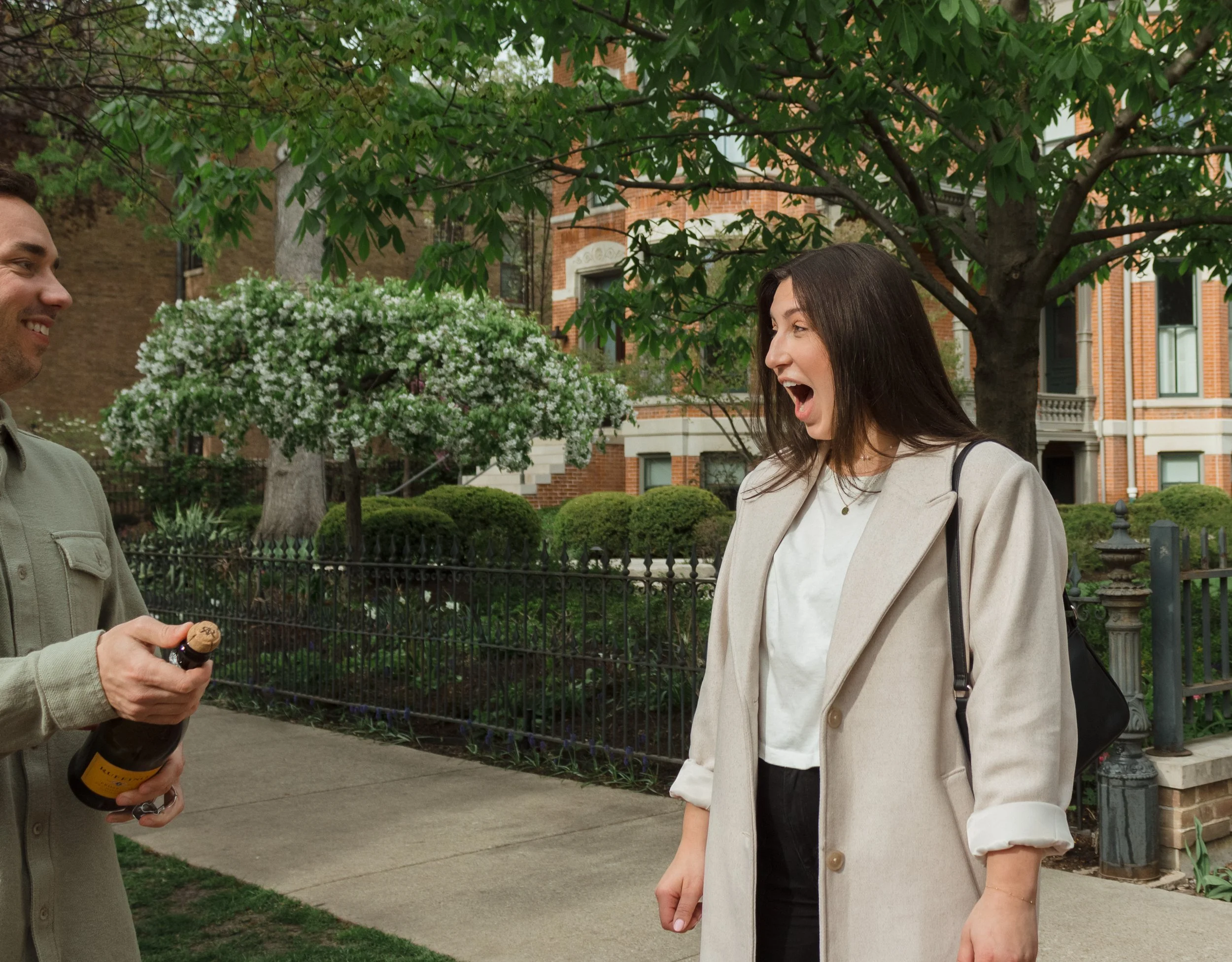 A woman with long dark hair, wearing a light-colored coat, is laughing with surprise as a man offers her a bottle of wine on a sidewalk in front of a fence and lush greenery, with brick houses in the background.