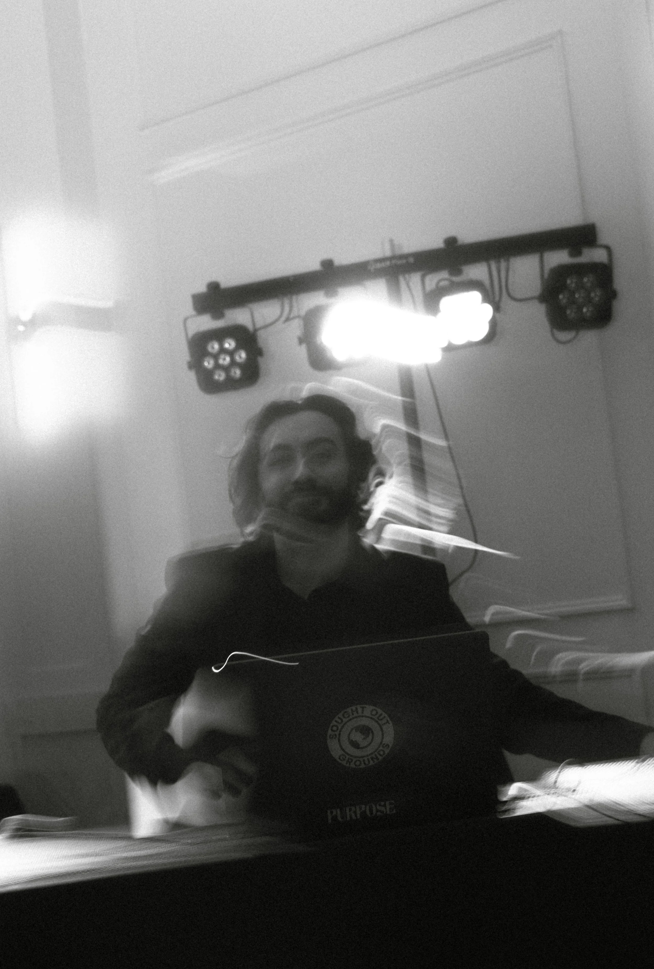 A black and white photo of a man with long hair and a beard, smiling, sitting at a table with a laptop that has a sticker reading "Sought Out Grounds Purpose." In the background, there are stage lights on a ceiling-mounted rig and a whiteboard or wal