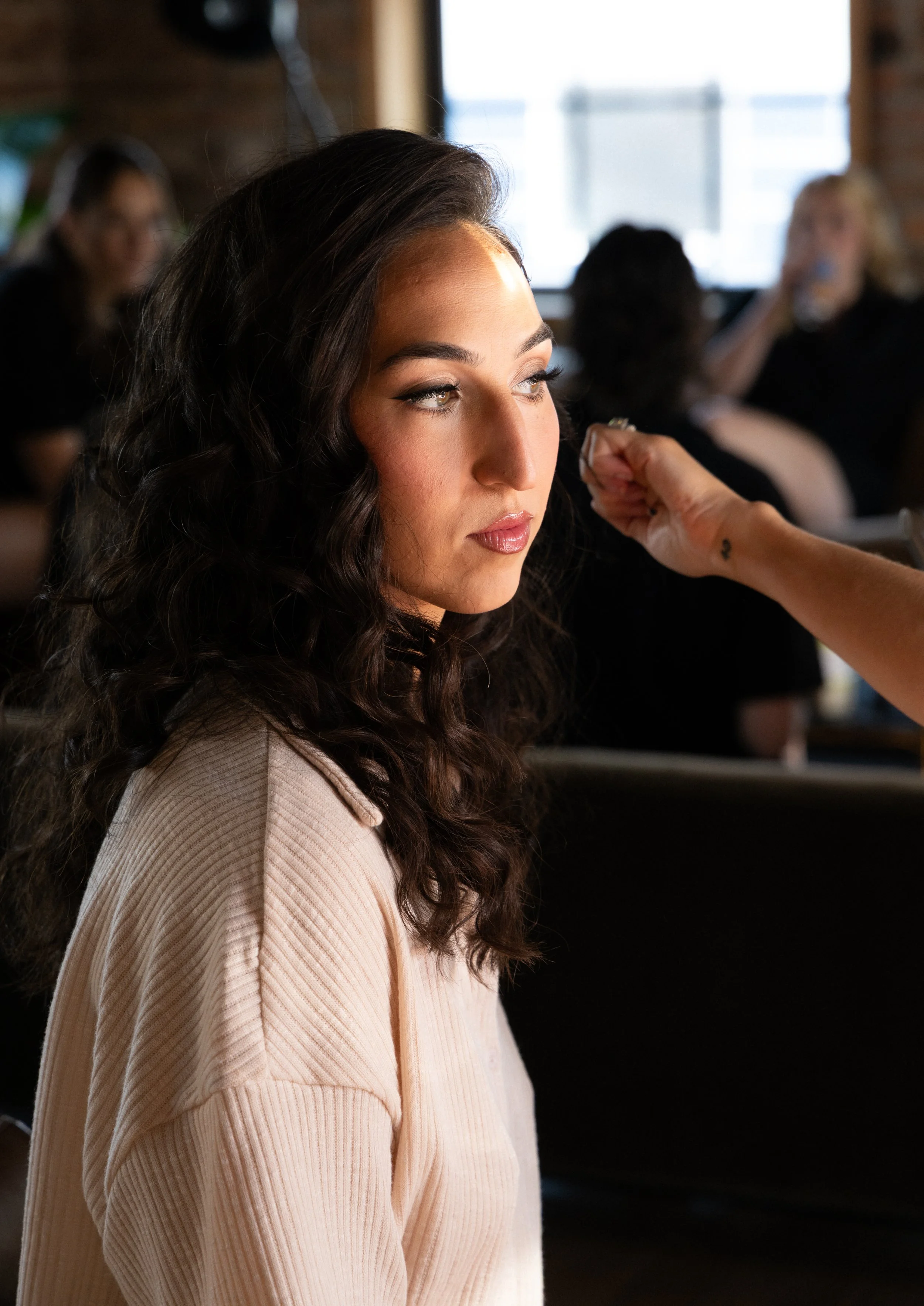 A woman with dark, curly hair and makeup sitting in a salon chair, receiving makeup application from a stylist's hand, while other women are seen in the background at a busy salon.