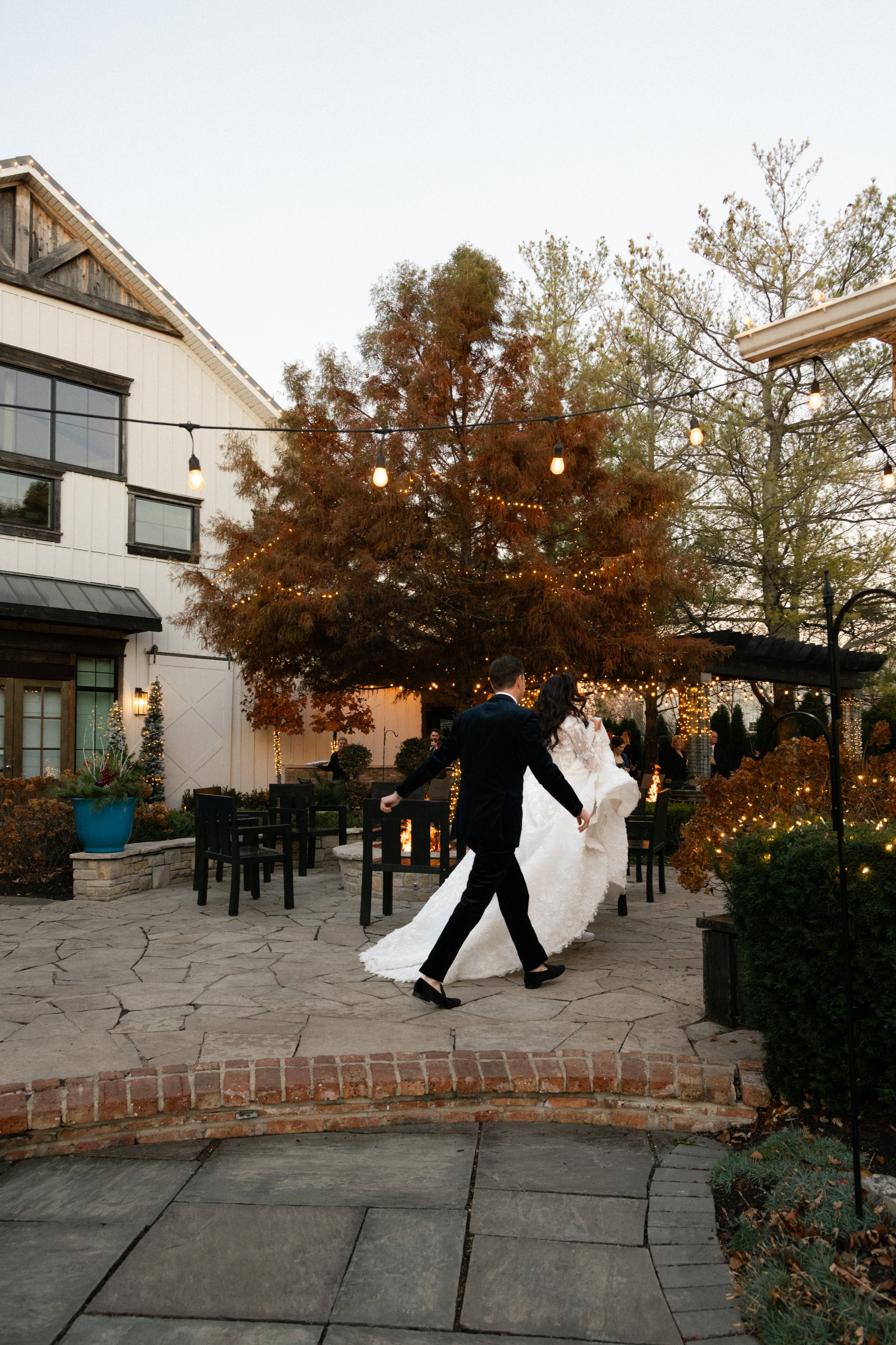 A bride and groom dancing outdoors during a wedding celebration at sunset, decorated with string lights, trees, and outdoor furniture.