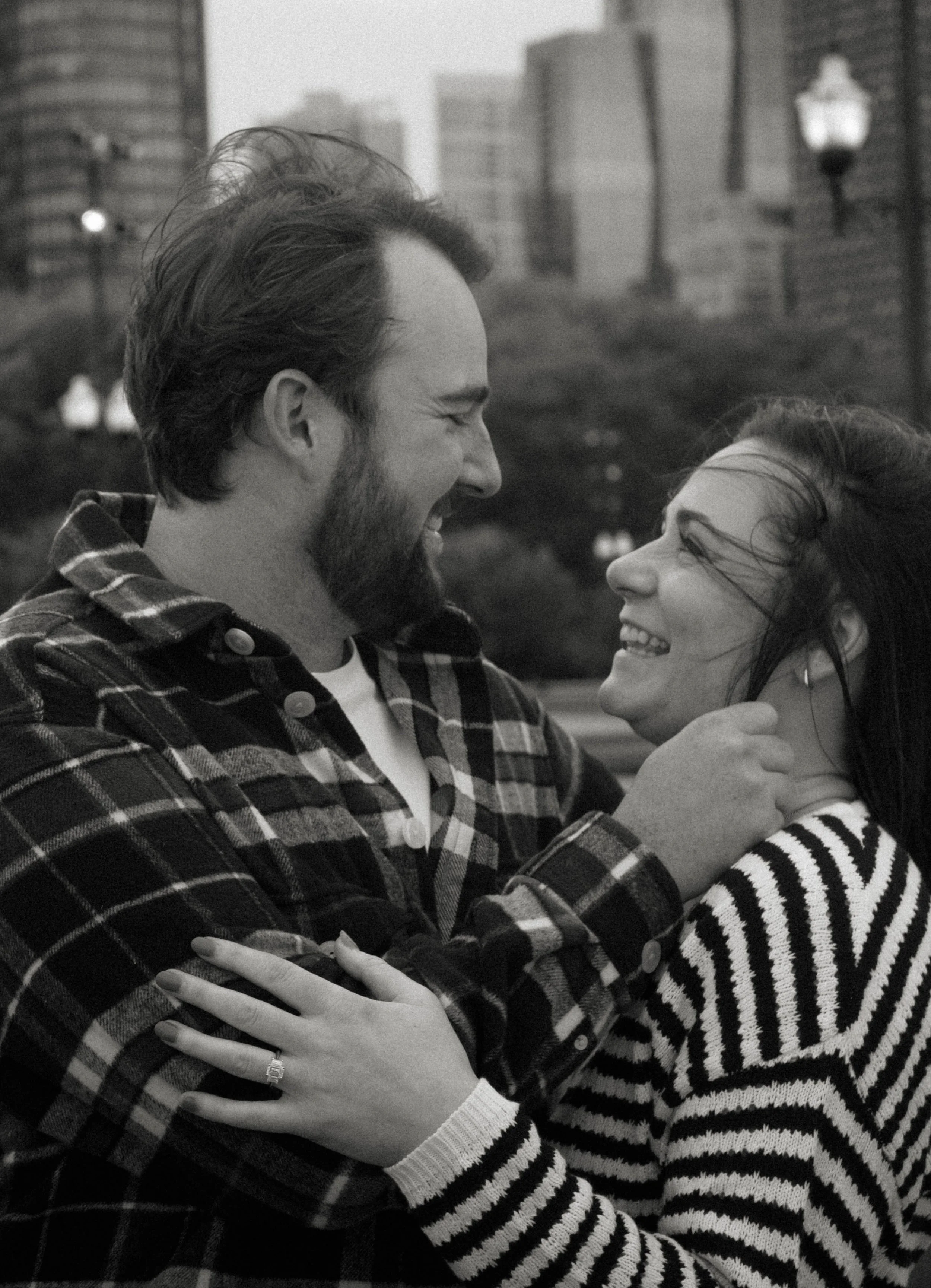 A happy couple smiling and embracing each other outdoors in an urban setting, with tall buildings and street lamps in the background.