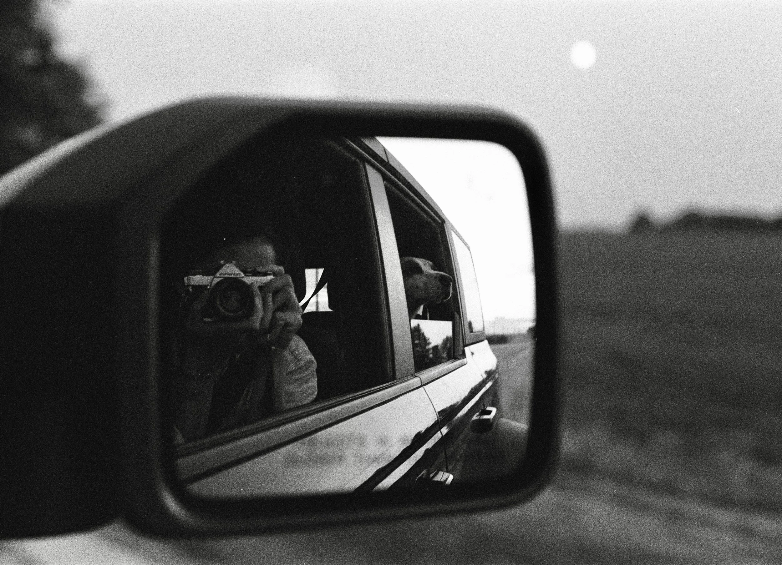 Black and white film photo of a person taking a picture with a camera in the rearview mirror of a car. The reflection shows a dog looking out of the vehicle's window and a landscape with a sunset or sunrise in the background.