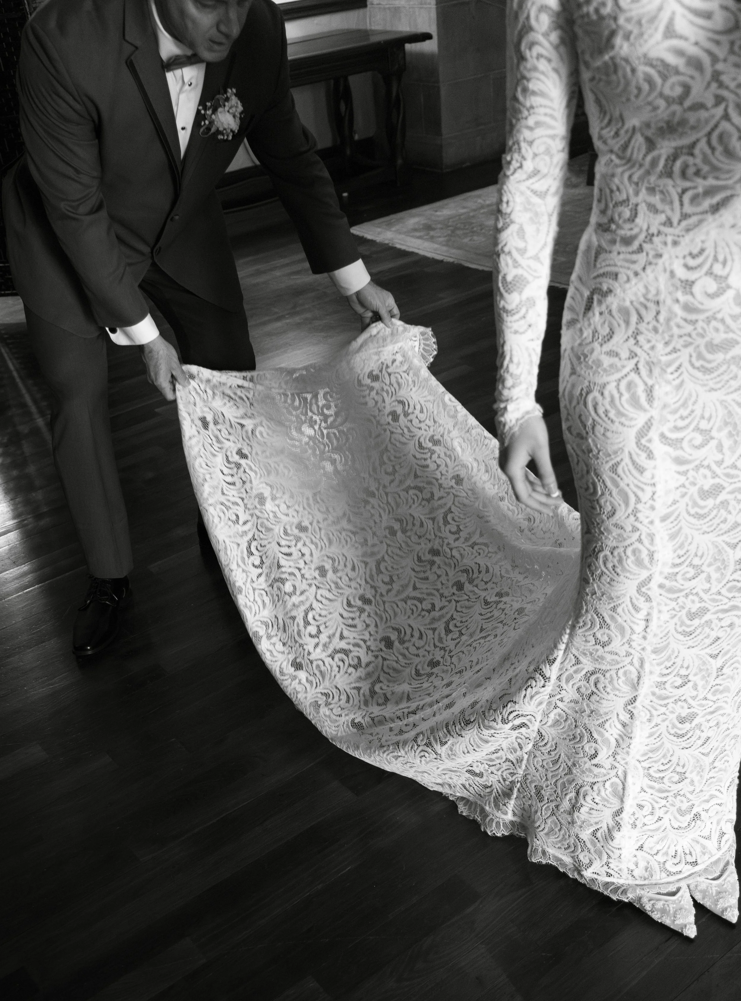A man in a tuxedo and a woman in a lace dress are handling a lace wedding train, preparing for a wedding or wedding photos.