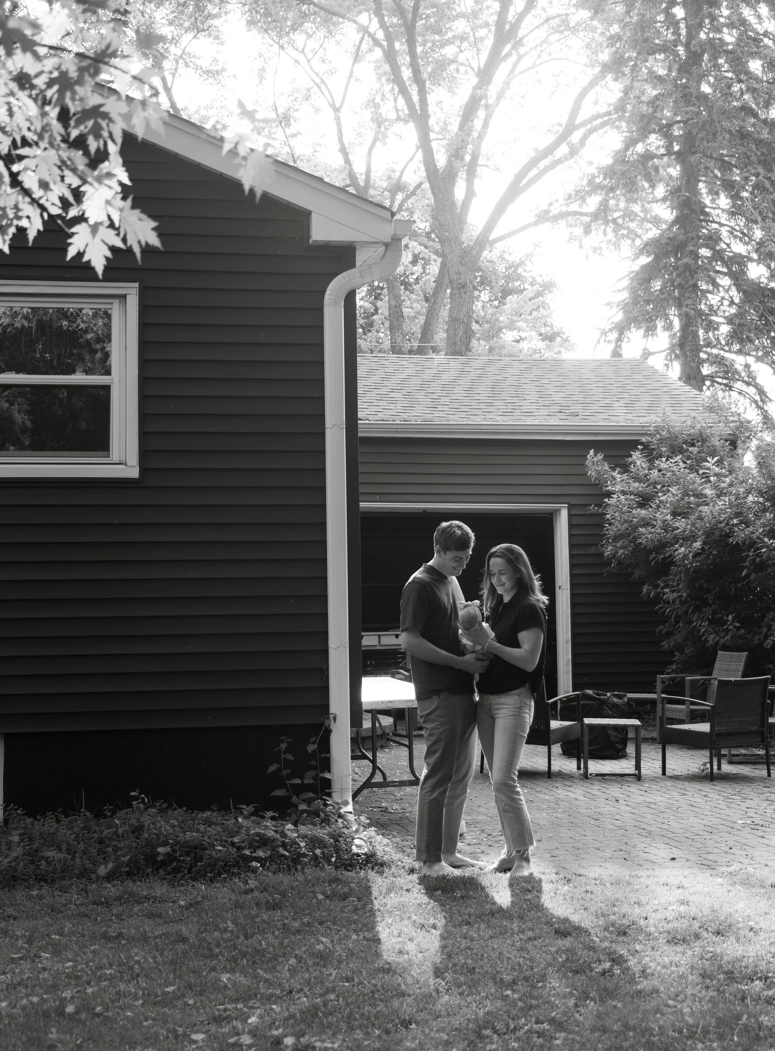 A couple standing outside their house holding a baby, with outdoor furniture and trees in the background, in black and white.