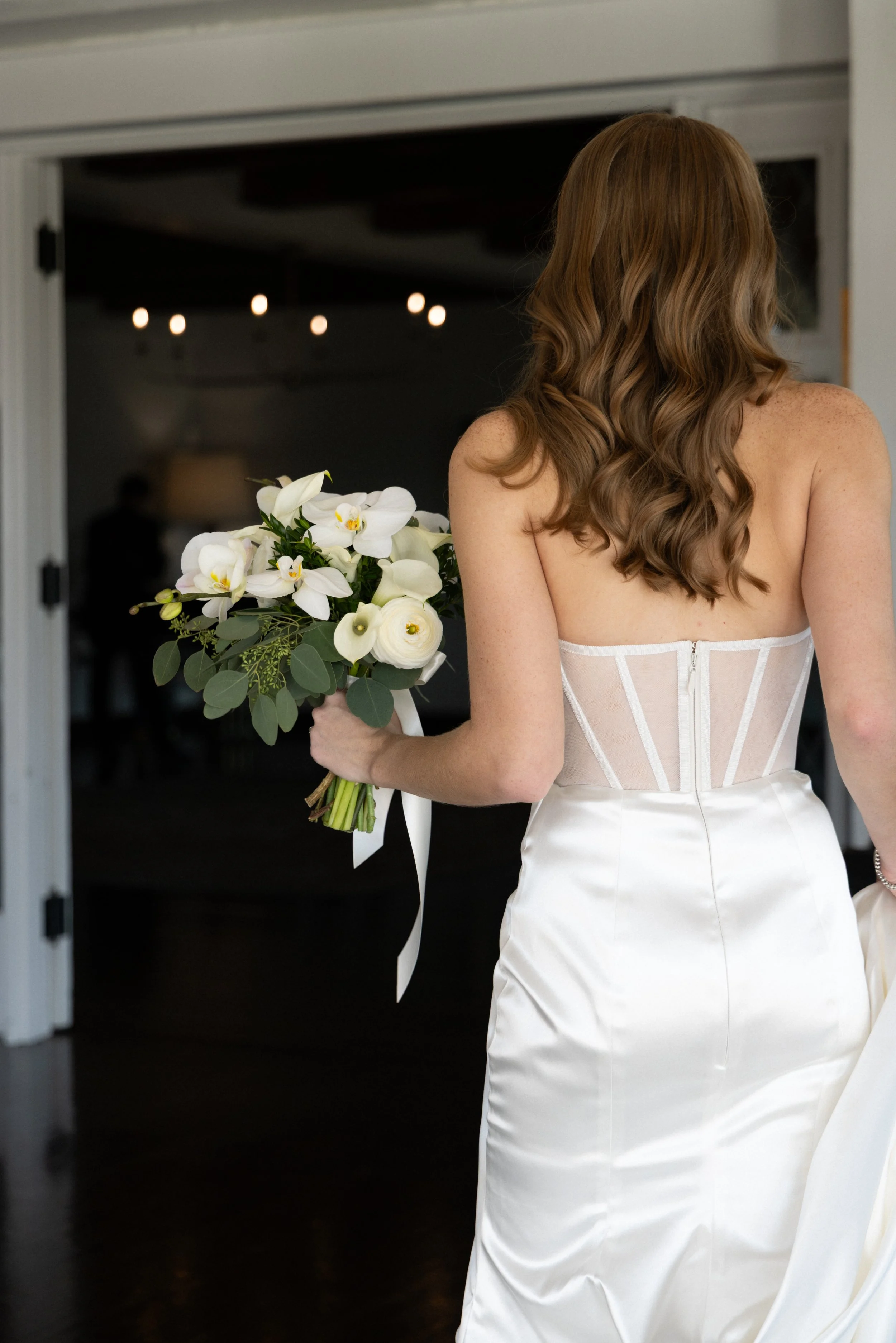A bride in a wedding dress holding a bouquet of white flowers, viewed from the back as she stands near an open doorway.