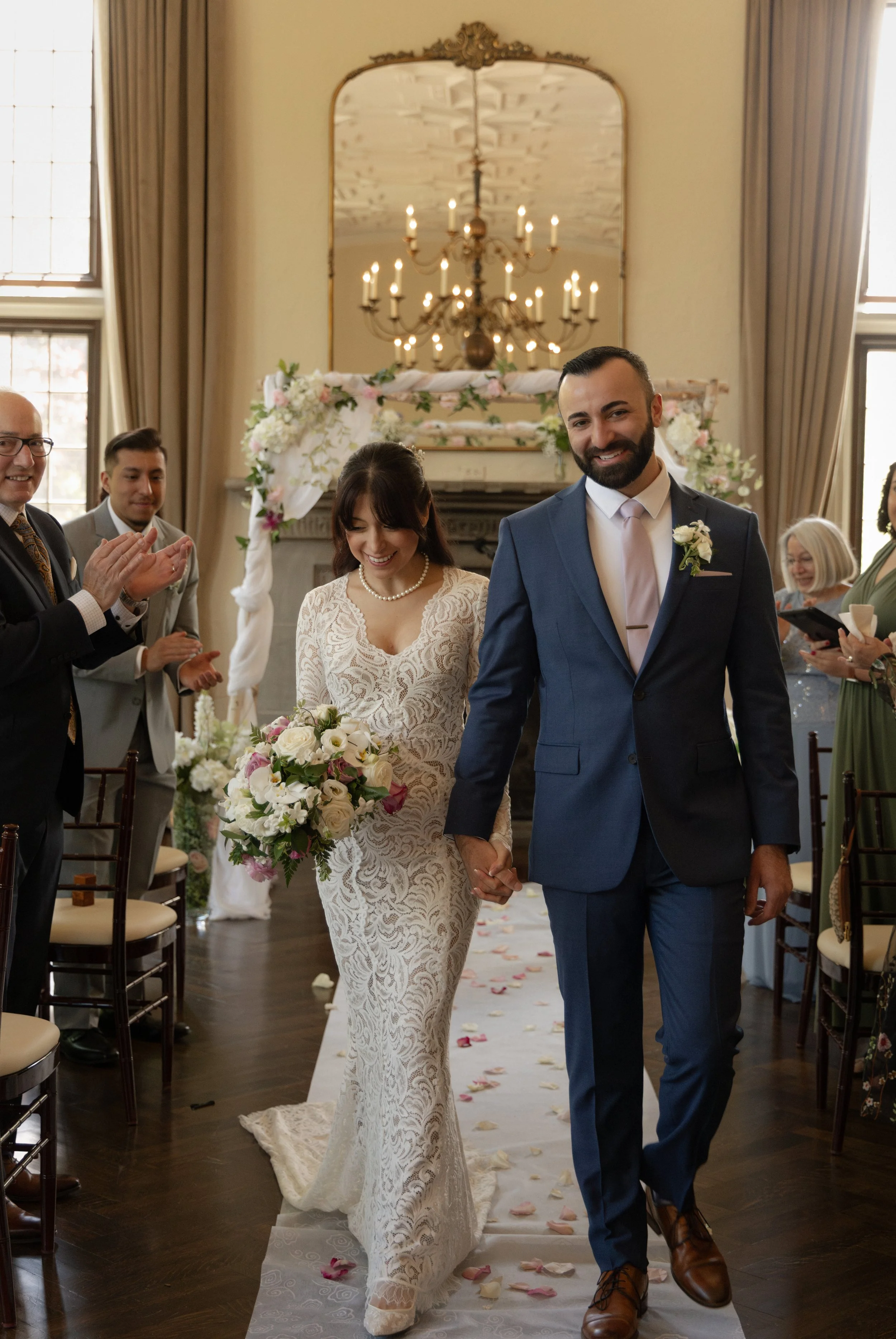 Bride and groom holding hands, walking down the aisle after their wedding ceremony, surrounded by clapping guests in a decorated indoor venue.
