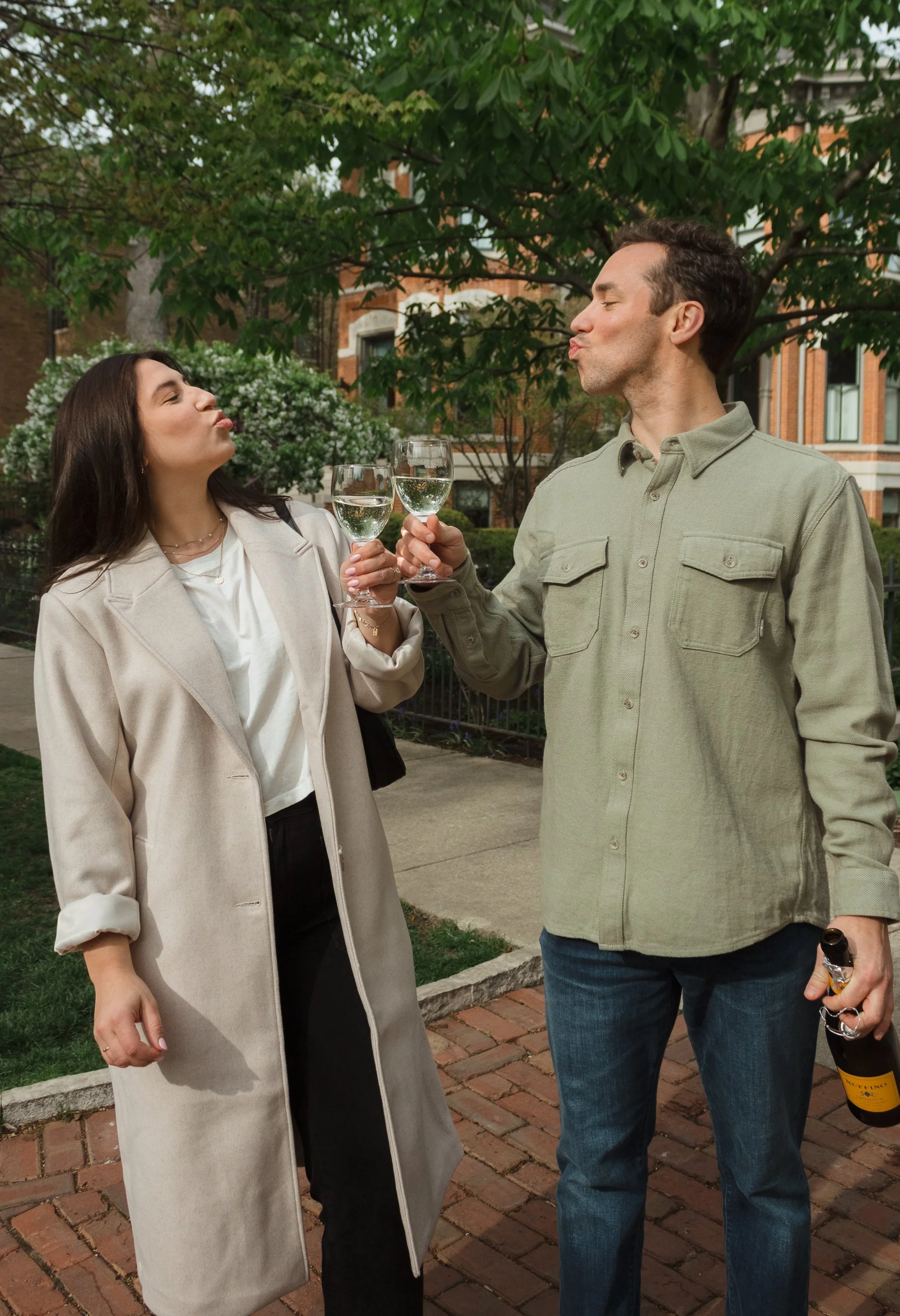 A man and woman celebrating with glasses of white wine outdoors on a brick sidewalk with trees and buildings in the background.