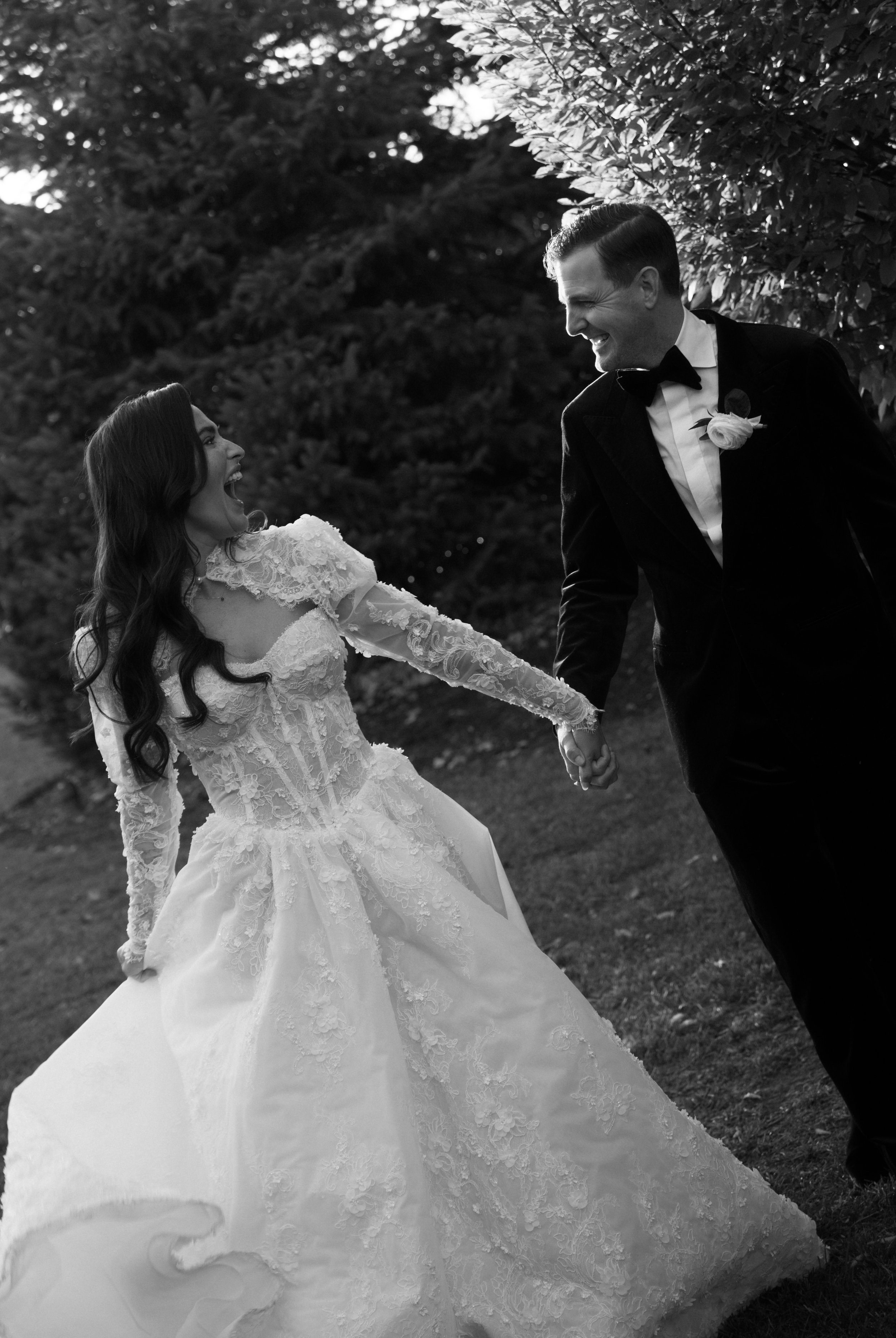 Black and white photo of a happy bride and groom holding hands and smiling outdoors during a wedding celebration.