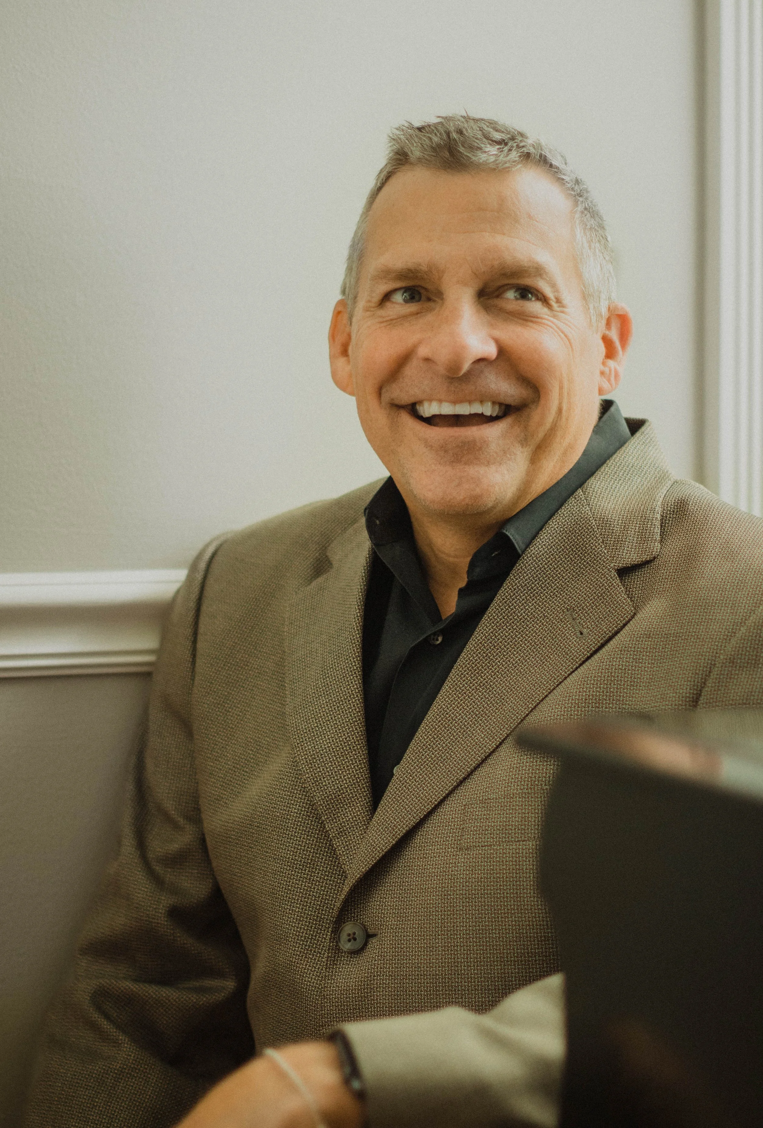 A middle-aged man with gray hair, smiling, wearing a tan blazer and black shirt, sitting against a light-colored wall.